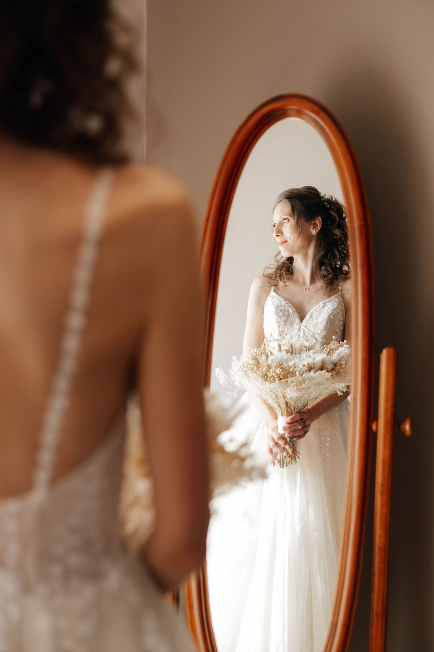 Bride in lace wedding dress holding dried floral bouquet reflected in oval wooden mirror.