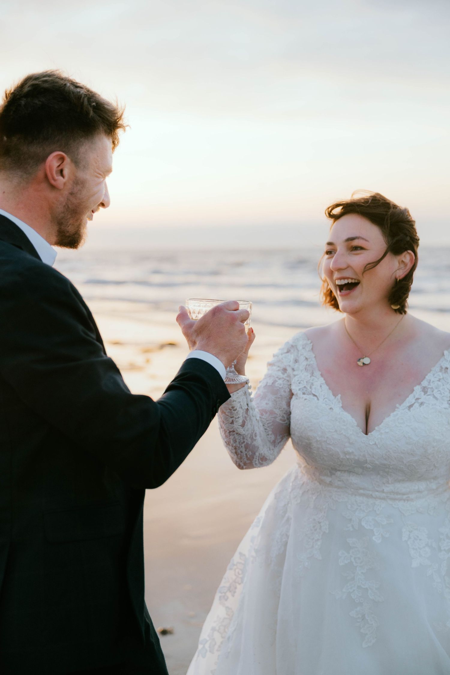 Bride and groom toasting with champagne on a beach at sunset, sharing a joyful laugh in wedding attire.