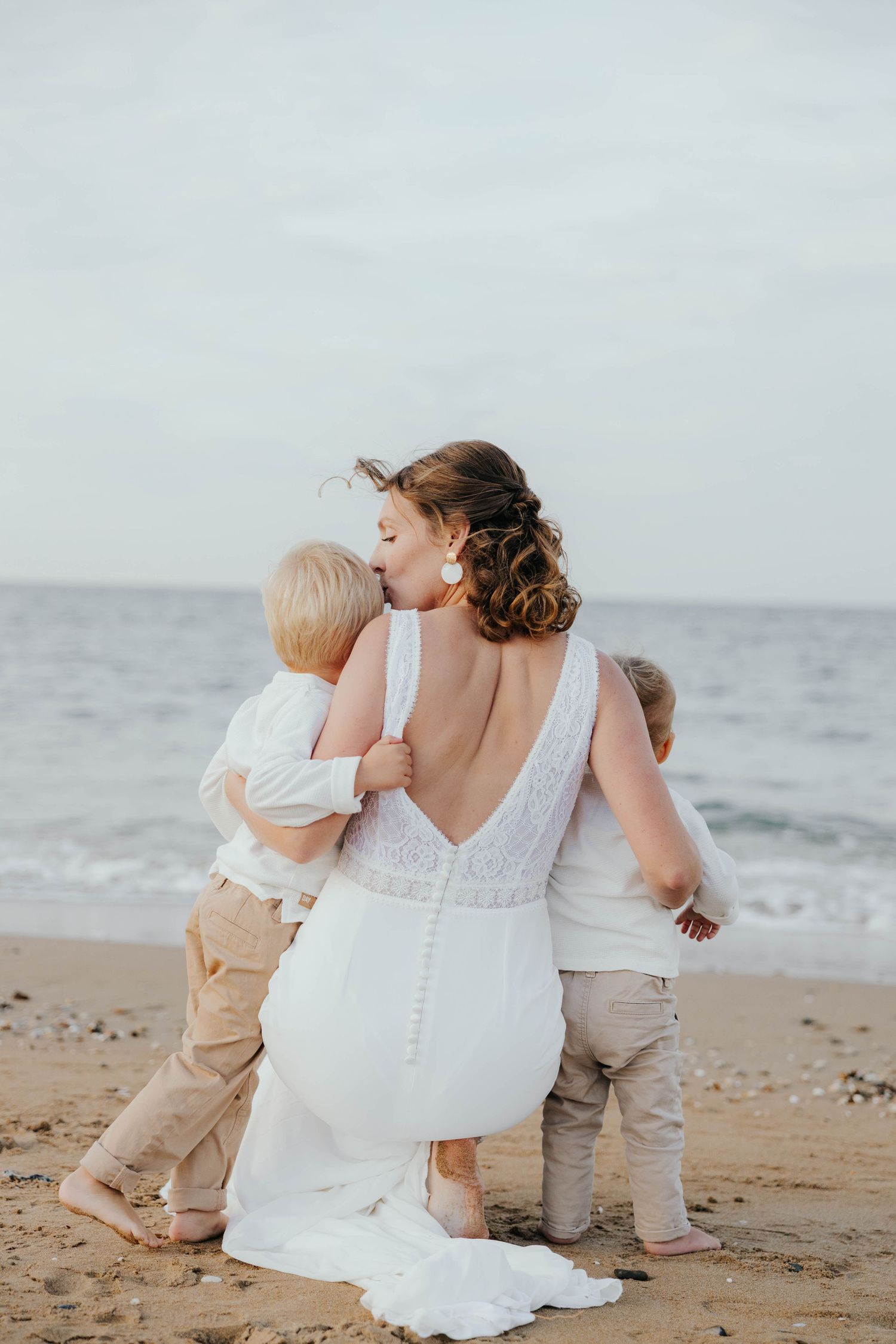 Bride in backless white gown embracing two young boys on a sandy beach with ocean waves behind her.
