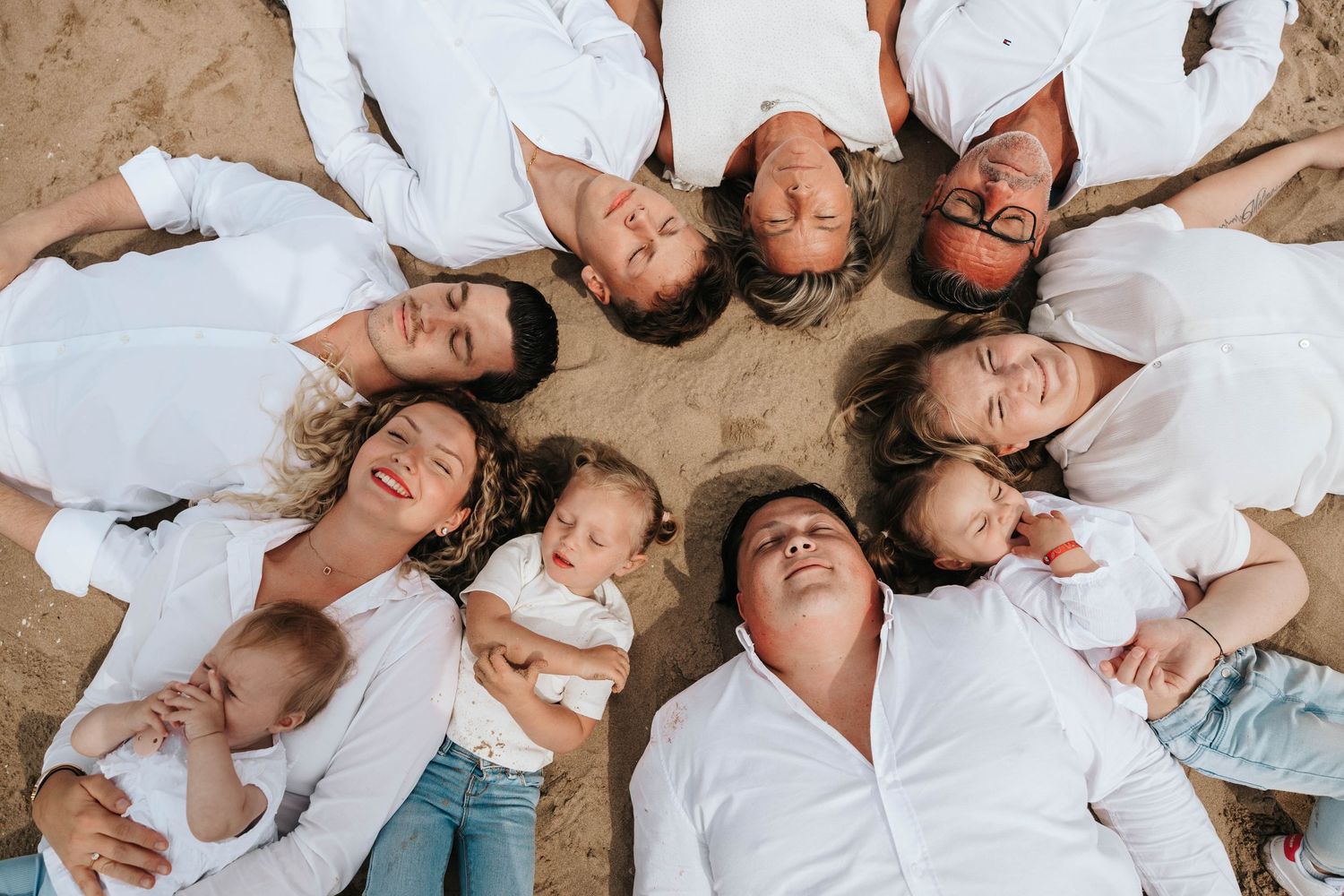 Aerial view of family in white shirts lying in a circle on sand with babies.