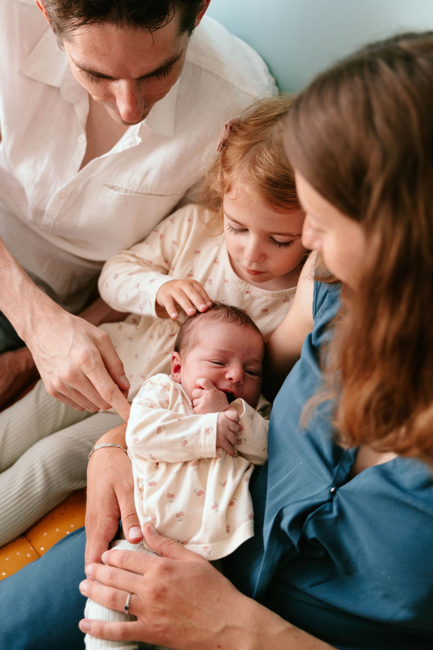 Parents and toddler sister lovingly gaze at and touch their sleeping newborn baby together.