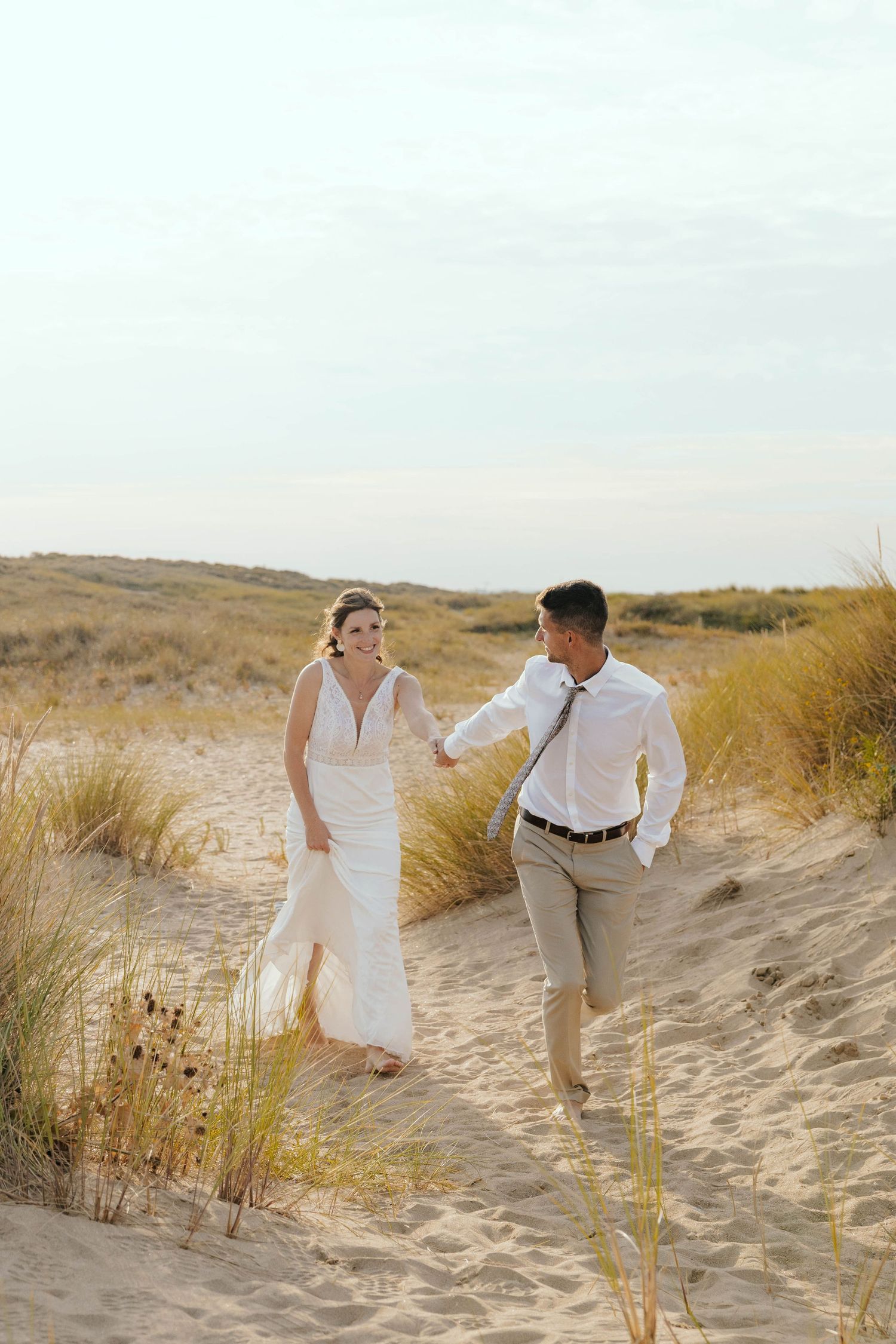 Couple running playfully through sandy beach dunes on wedding day.