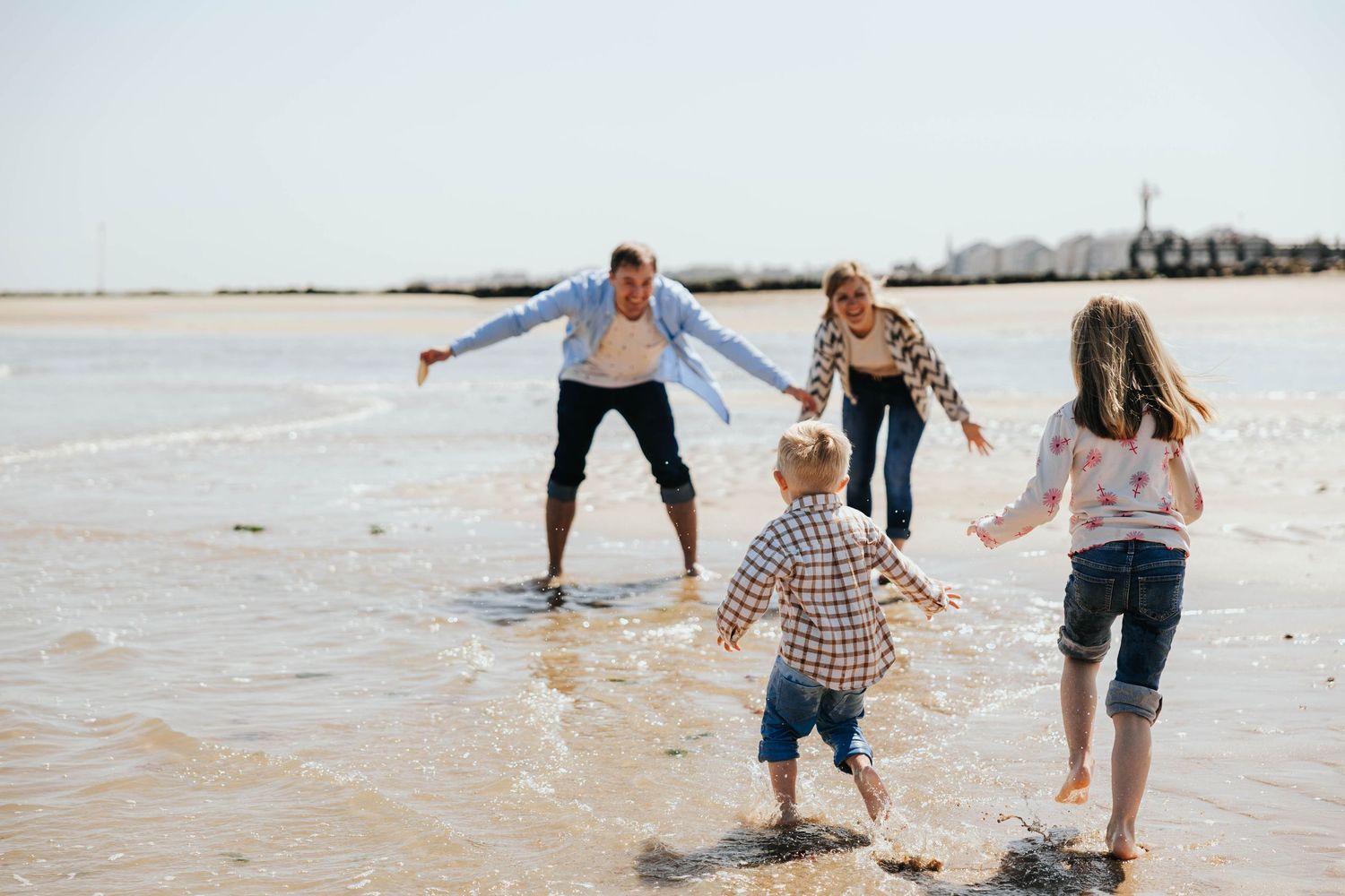 A cheerful family of four plays together in shallow ocean waves on a bright sunny beach day.