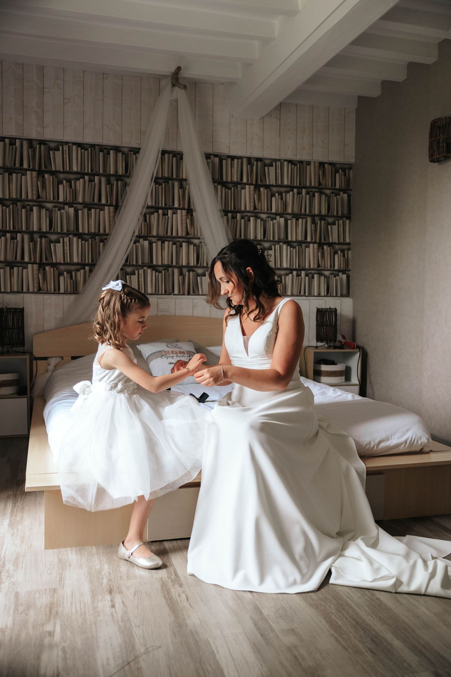 Bride in white gown sitting on bed sharing a tender moment with young flower girl in white dress.