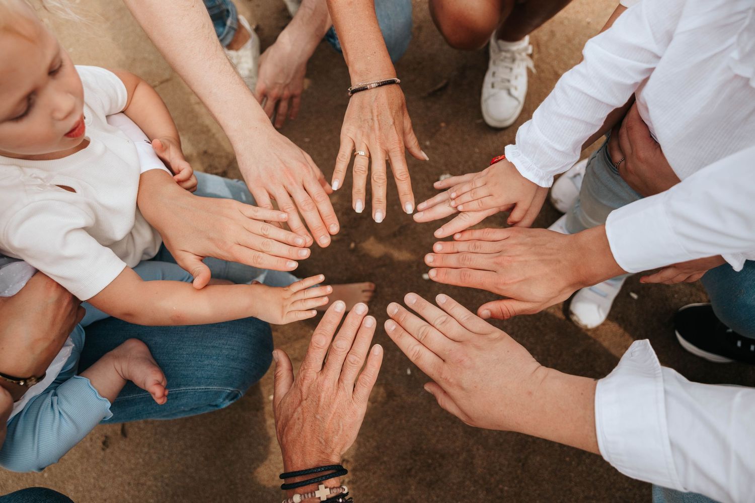 Family members placing hands together in a circle on sandy ground outdoors.