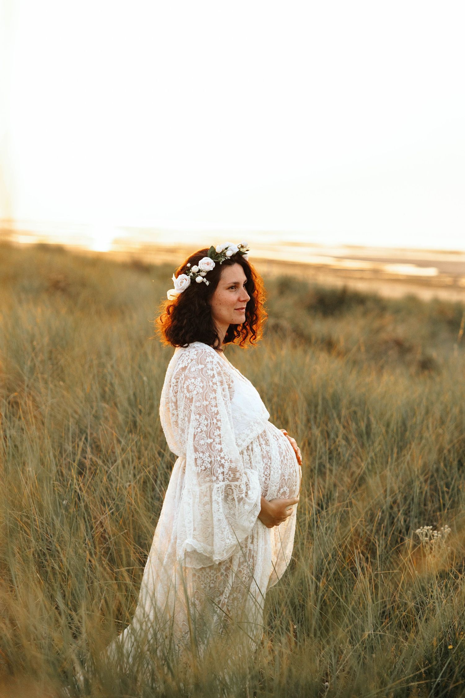 Pregnant woman in white lace dress with flower crown standing in golden field at sunset.