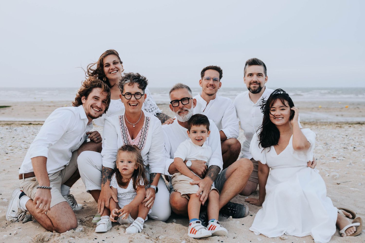 Multi-generational family in white outfits posing together on a sandy beach with ocean in background.