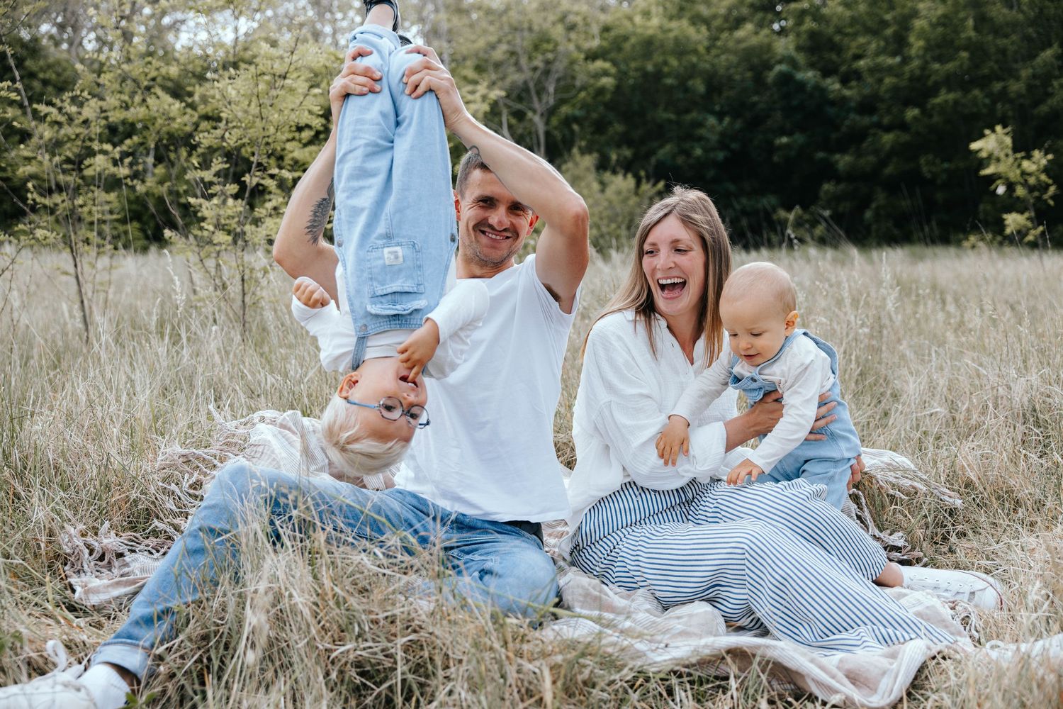 Happy family sitting in a grassy field, parents playing with two young children outdoors on a sunny day.