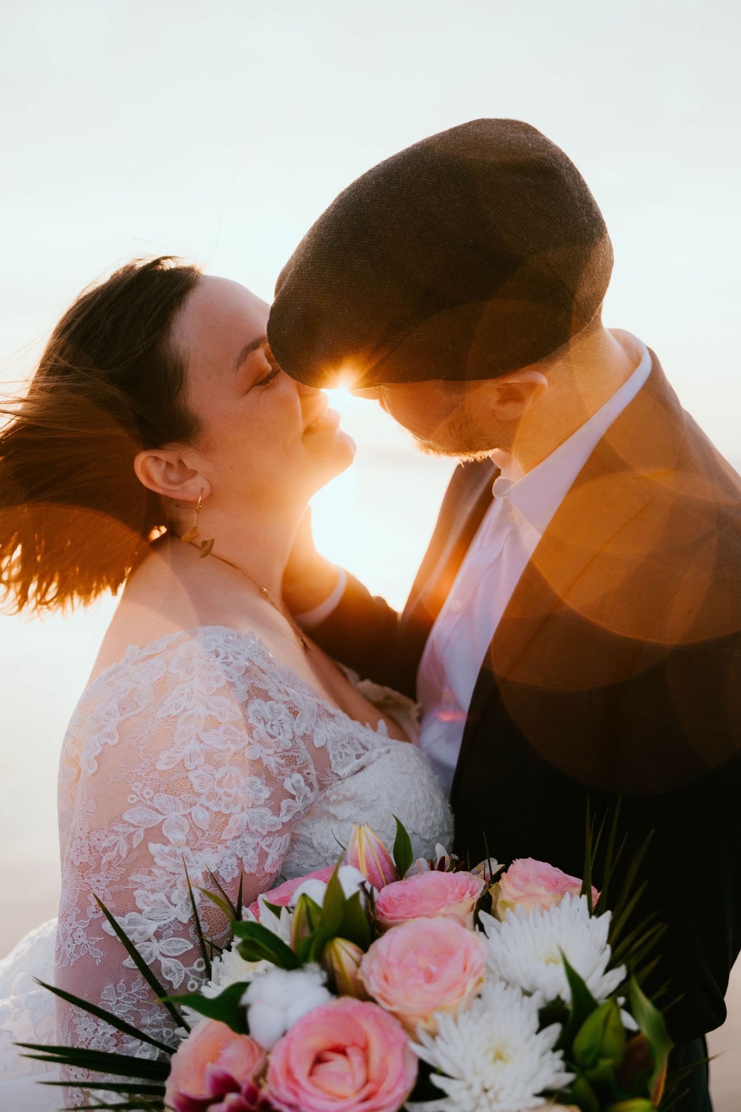 Bride and groom share a romantic sunset moment, bride holding a pink and white bouquet in a lace wedding dress.