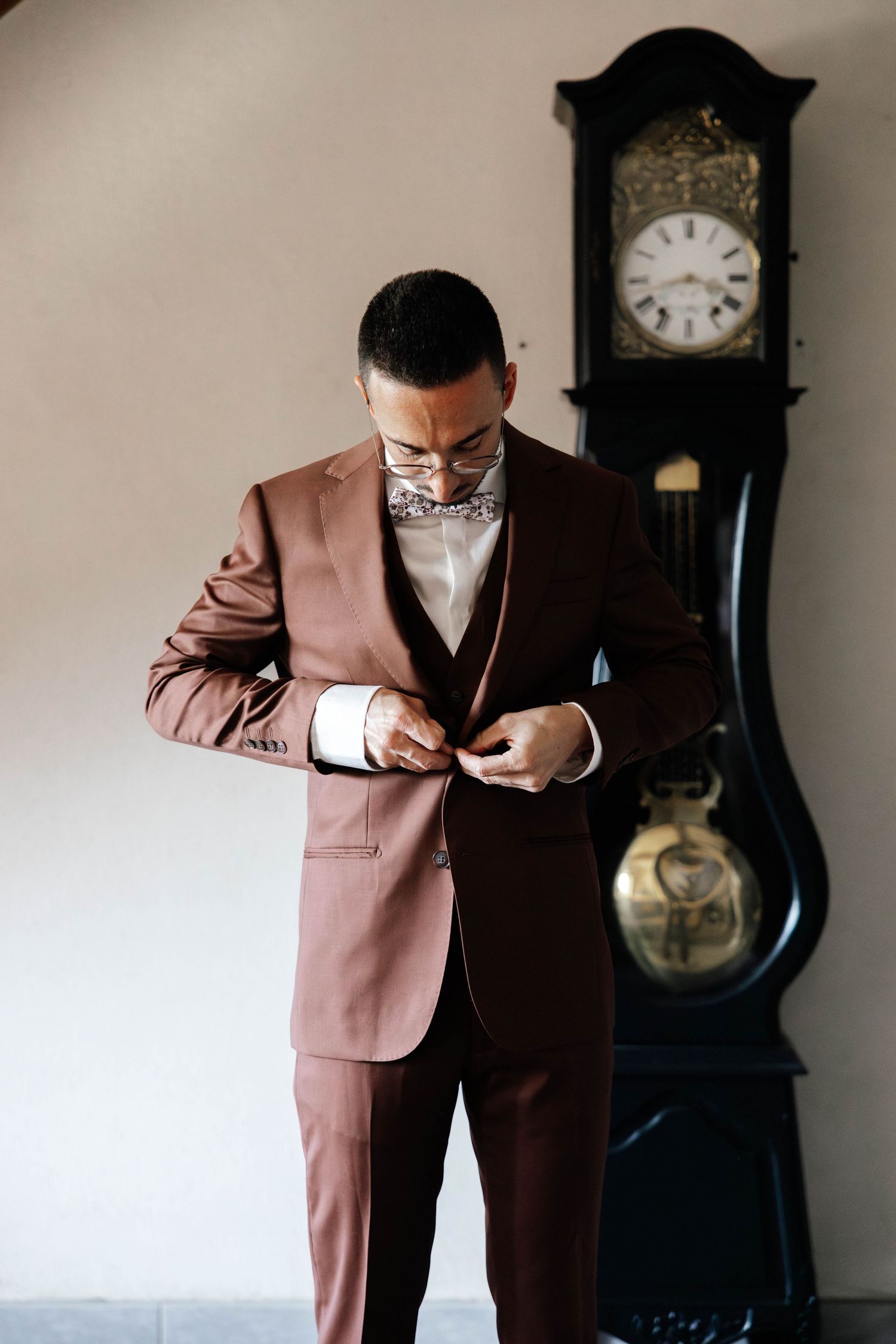 A groom in a brown satin suit adjusts his jacket button beside a vintage grandfather clock.