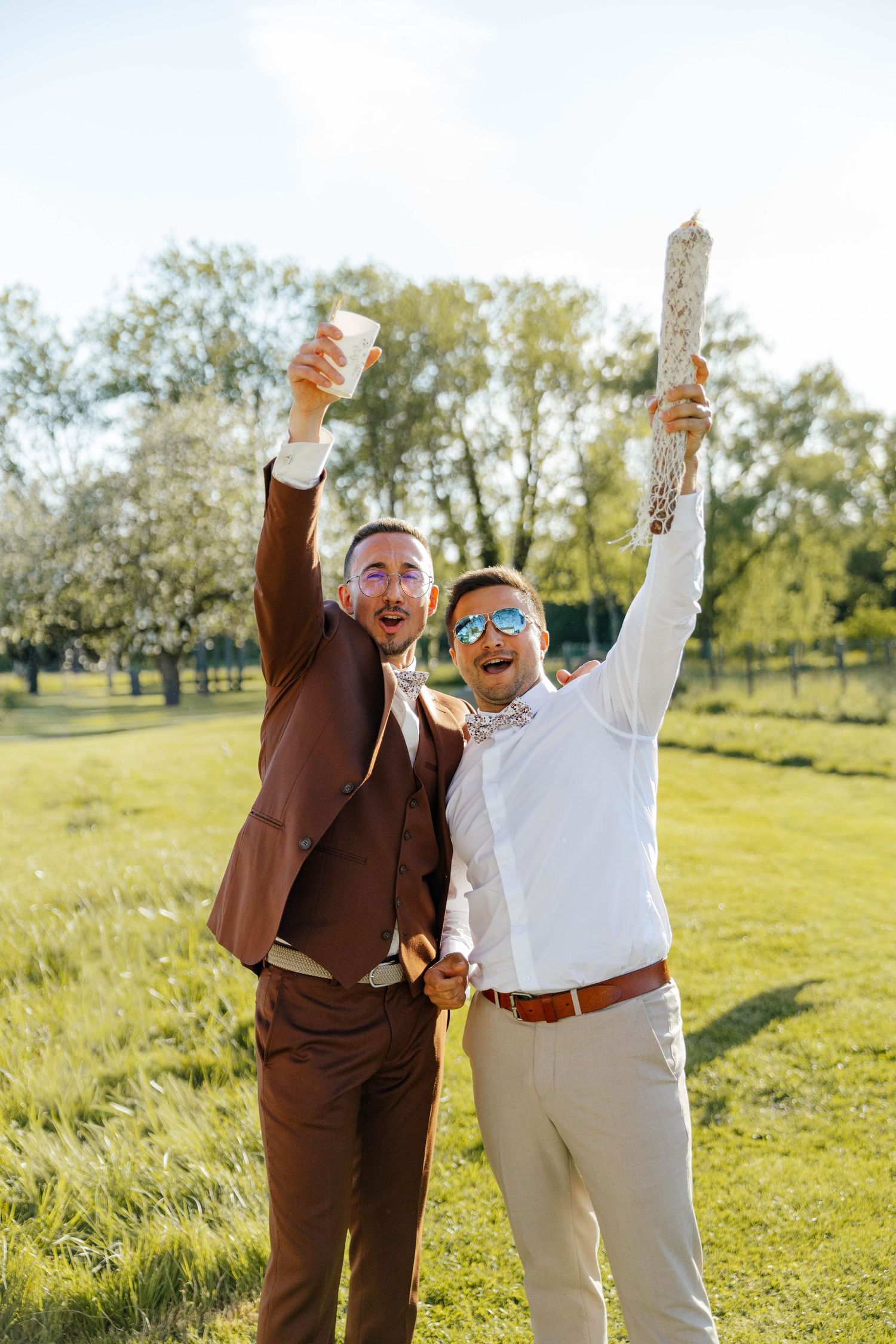Two groomsmen celebrating outdoors raising drinks and laughing in sunny garden setting.