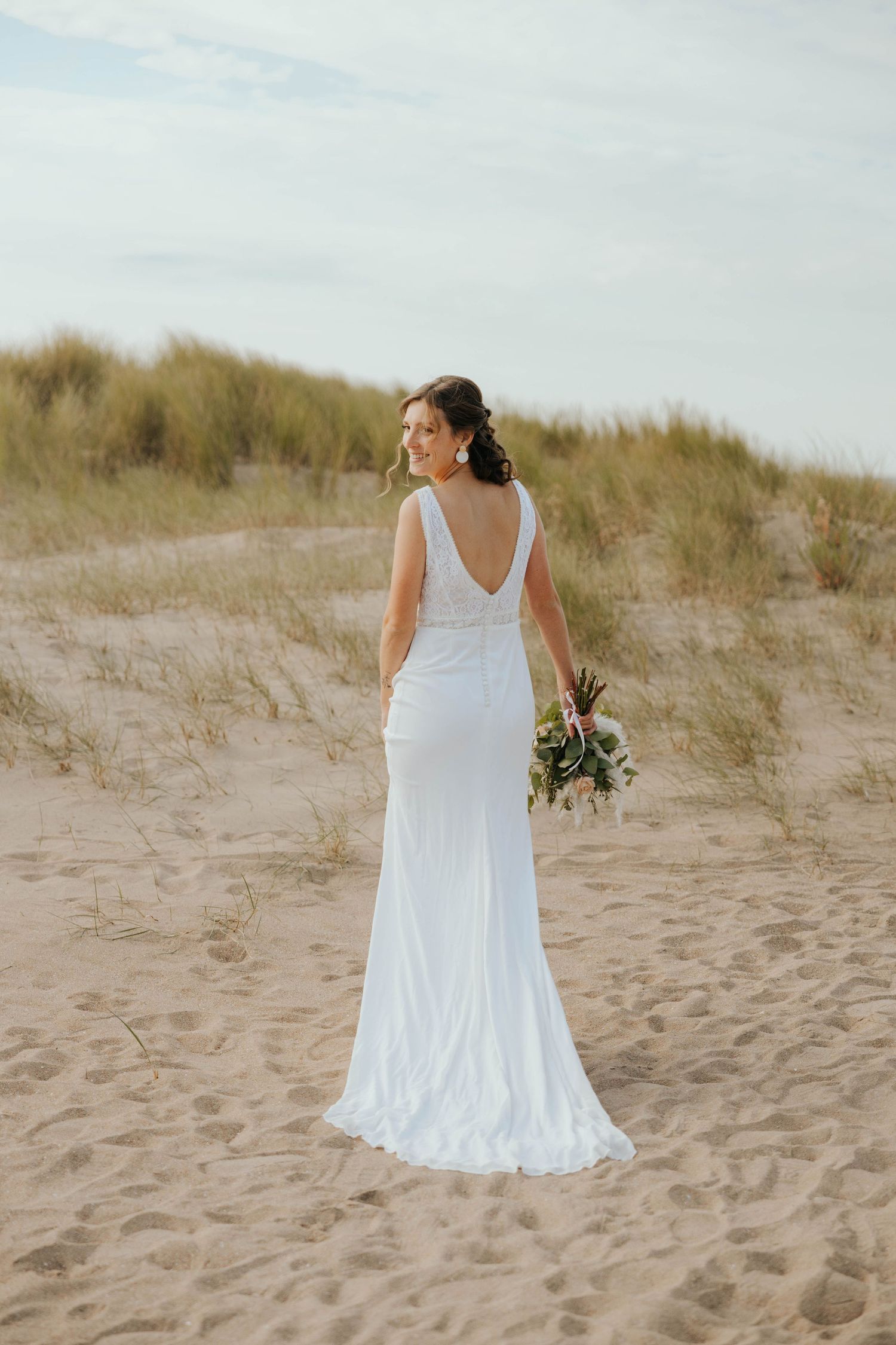 Bride in white backless gown holding bouquet on sandy beach.