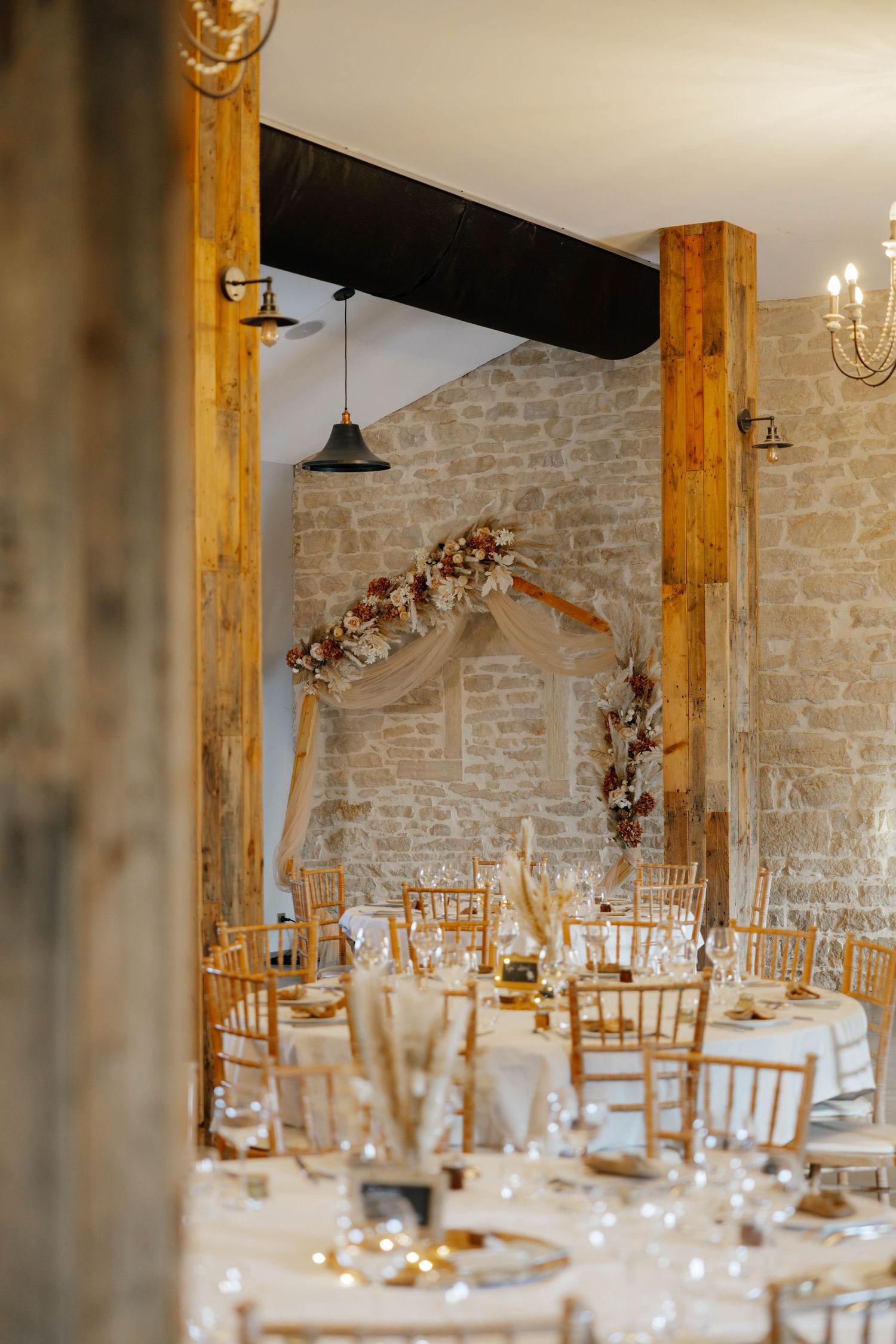 Rustic barn wedding reception with wooden beams, floral arch, and gold Chiavari chairs.