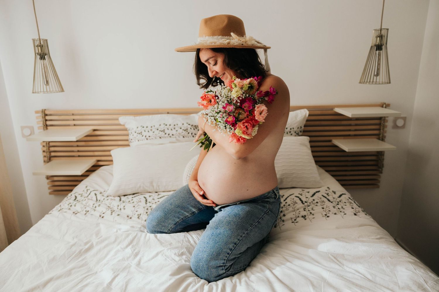 Pregnant woman in hat and jeans holds colorful flower bouquet while sitting on white bed.