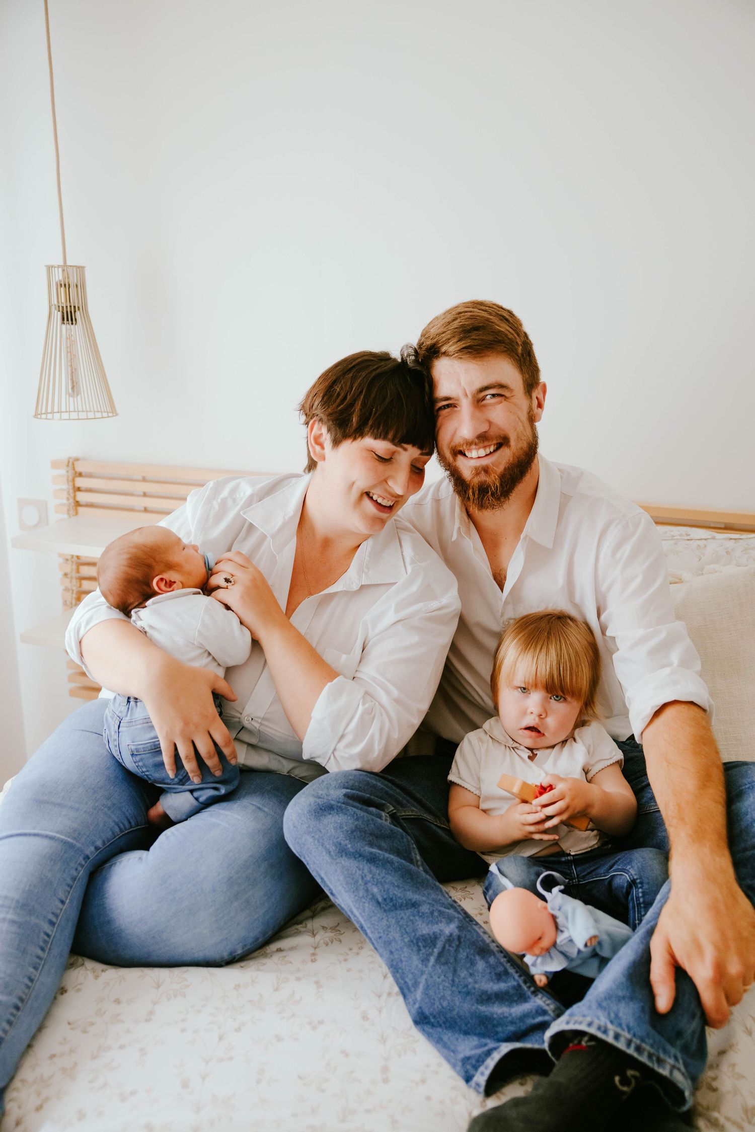 Smiling family with newborn and toddler sitting together on white bed in cozy bedroom.