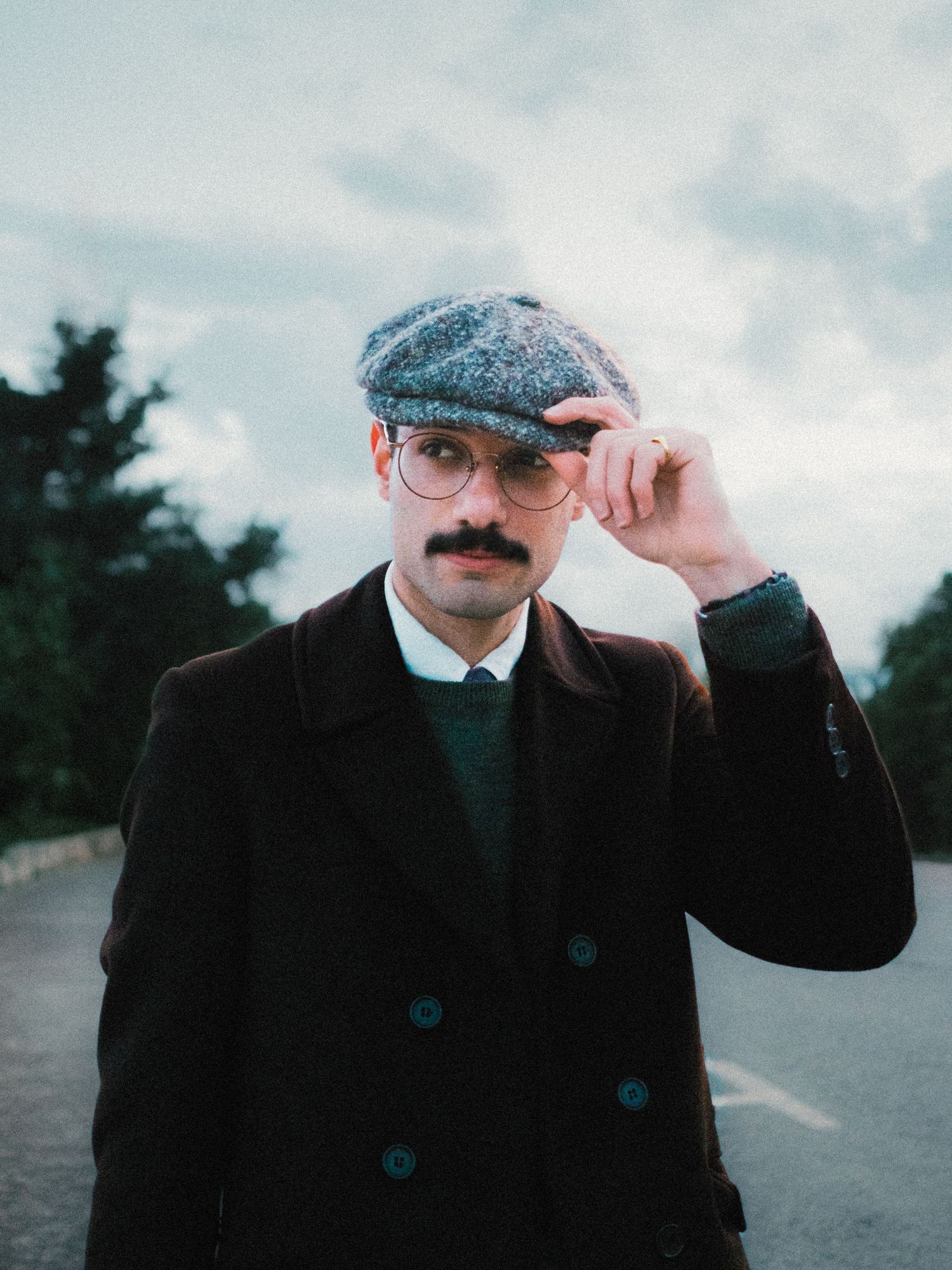 Retro-styled portrait of someone wearing a flat cap, glasses, and dark coat on an overcast day.