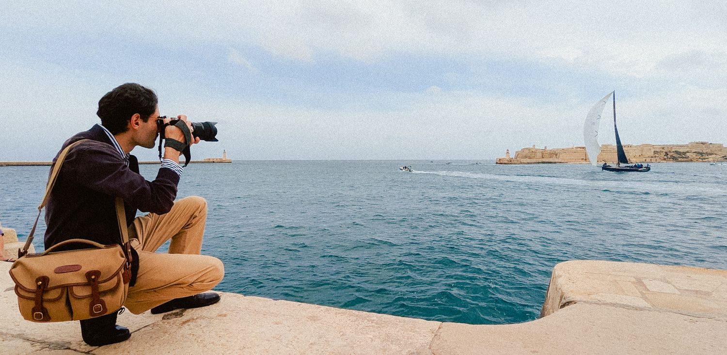 Photographer crouching on Mediterranean coast with camera photographing sailboat on turquoise waters near rocky islands.