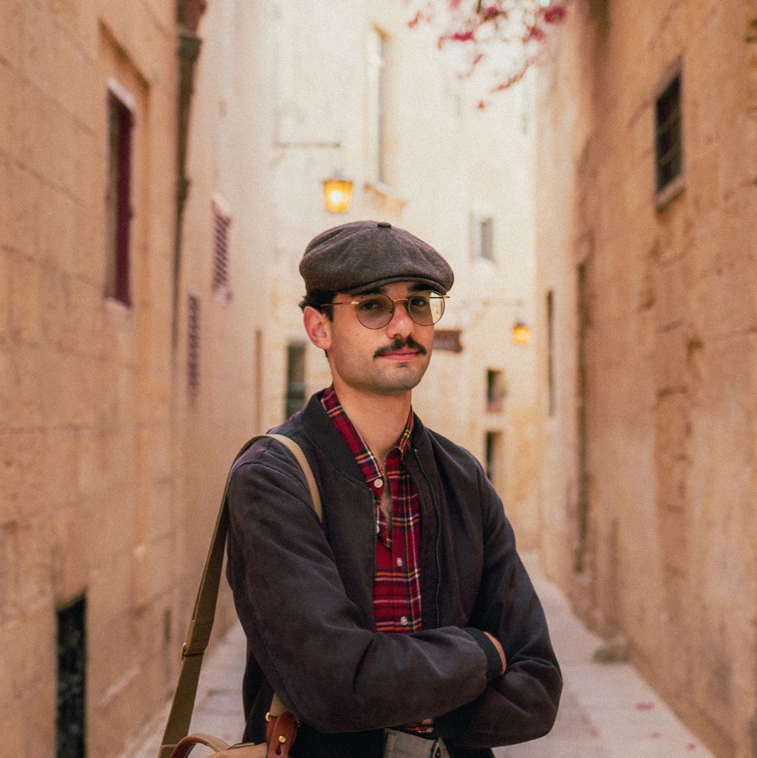 Person wearing a flat cap and dark jacket posing in a narrow beige stone alleyway of a Mediterranean town.