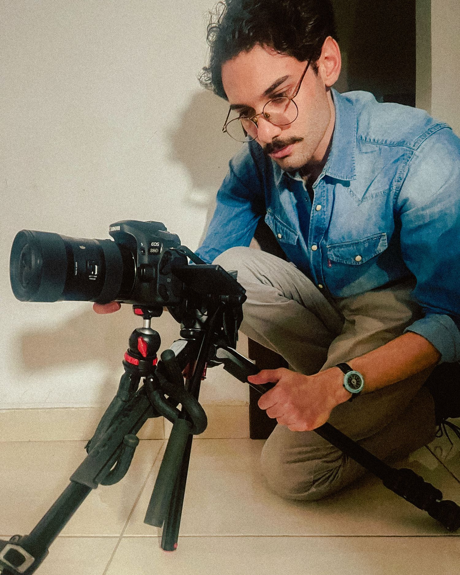 A photographer in denim shirt and khakis crouches beside a camera mounted on a professional tripod setup.