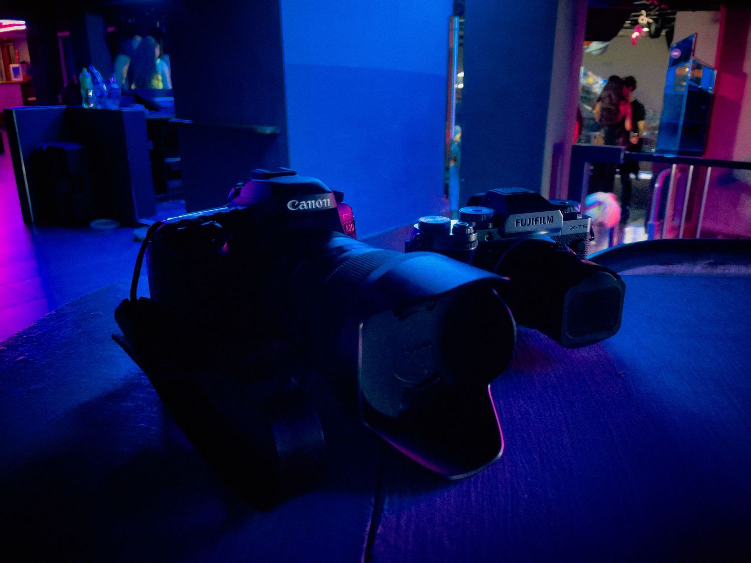 Two Canon DSLR cameras with lenses sit in a dimly lit room with purple and blue ambient lighting.