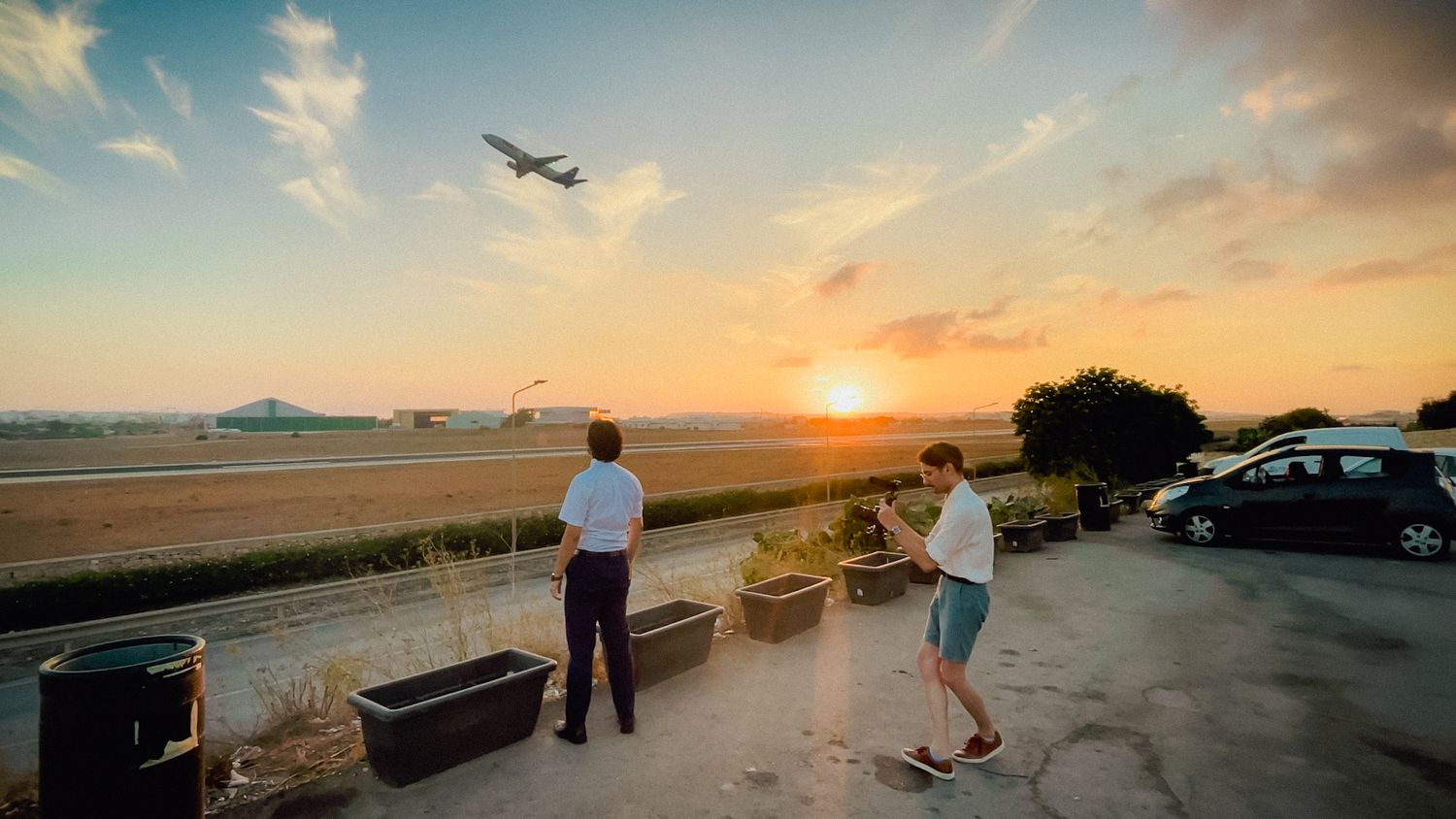 Two people watch a commercial airplane take off during golden sunset at an airport observation area.