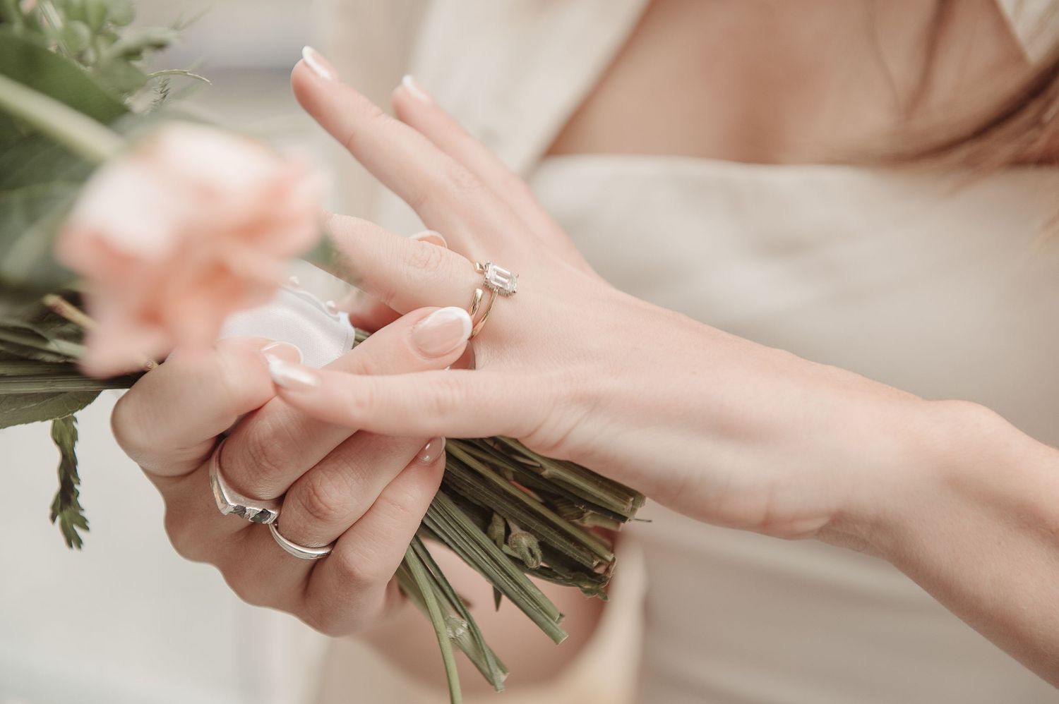 Close-up of a bride placing a diamond engagement ring on her finger while holding a soft pink floral bouquet.