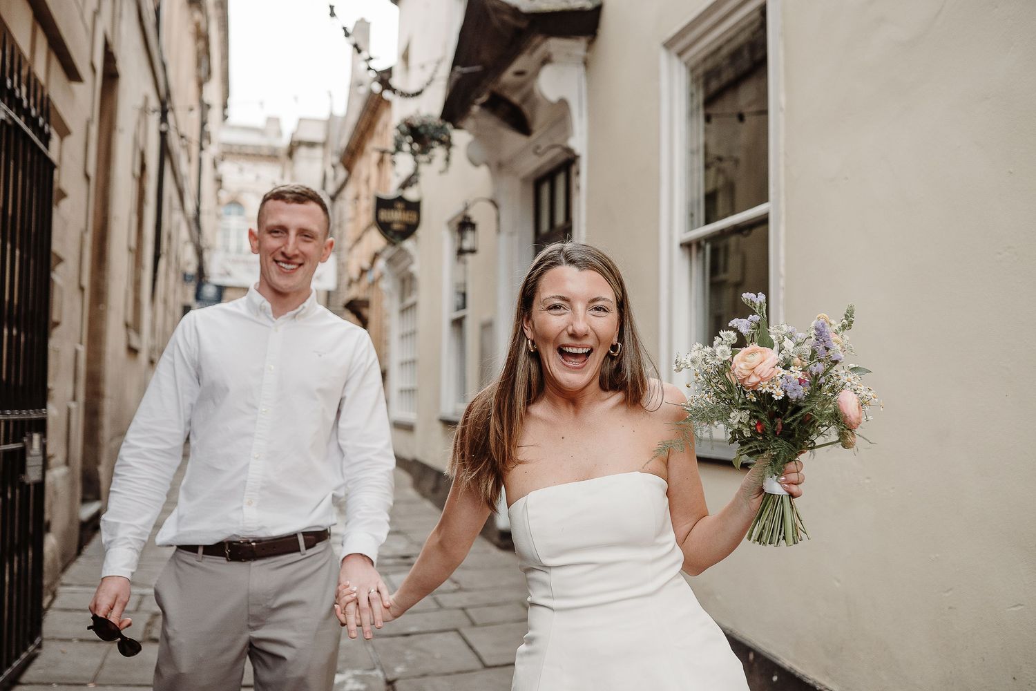 Joyful bride holding pastel bouquet walks cobblestone street with groom in white shirt on sunny wedding day.