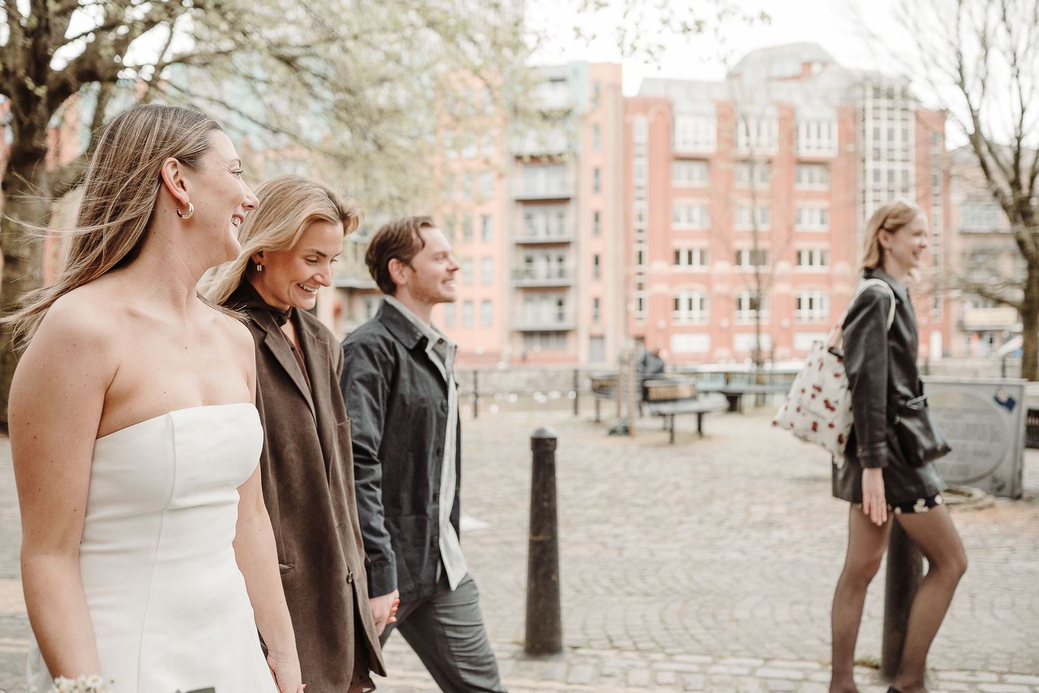 Four stylish young people walking on a cobblestone street in an urban setting with brick buildings in the background.