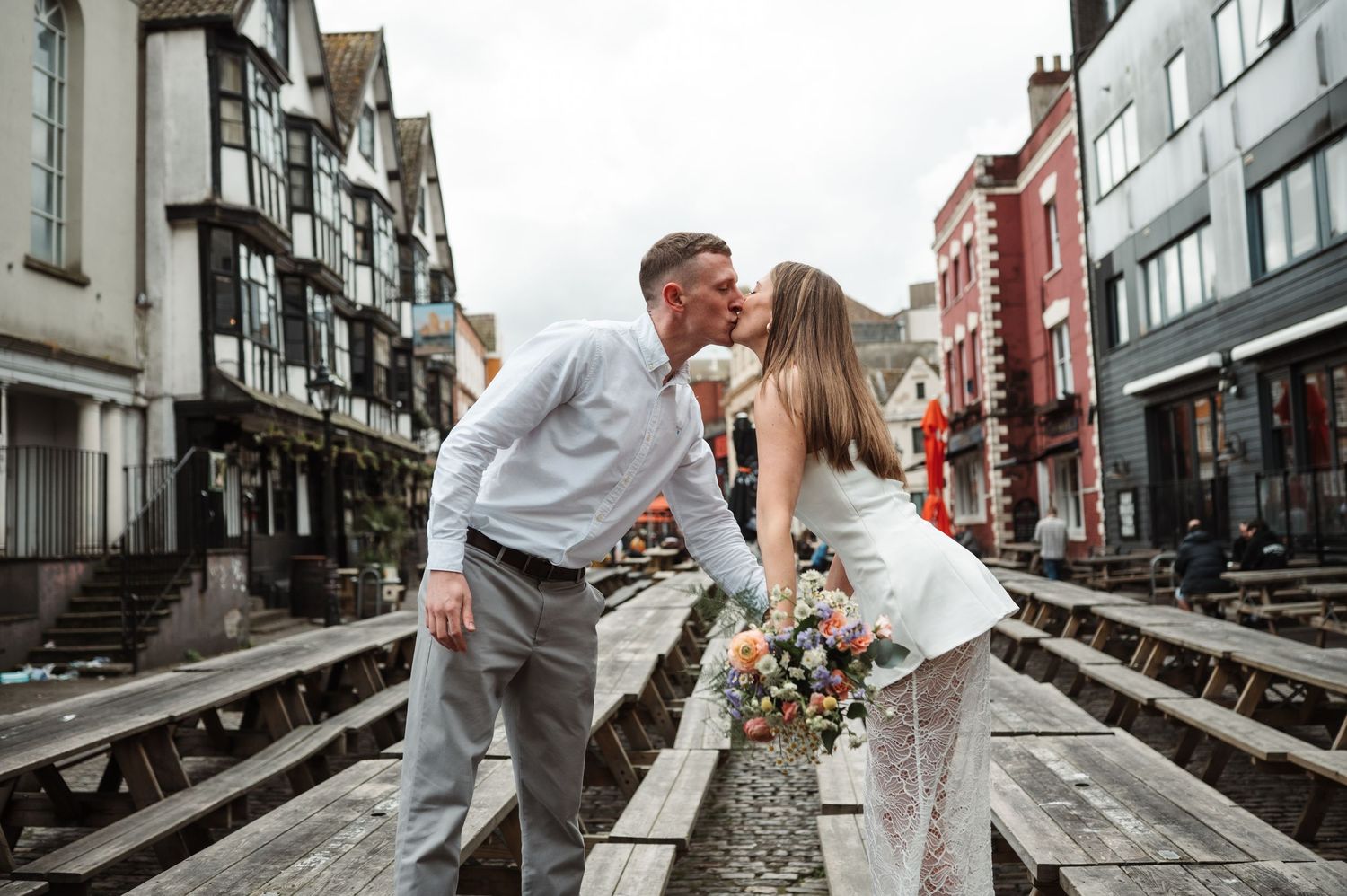 Newlywed couple kissing on wooden boardwalk with colorful bouquet, historic Tudor buildings in background.