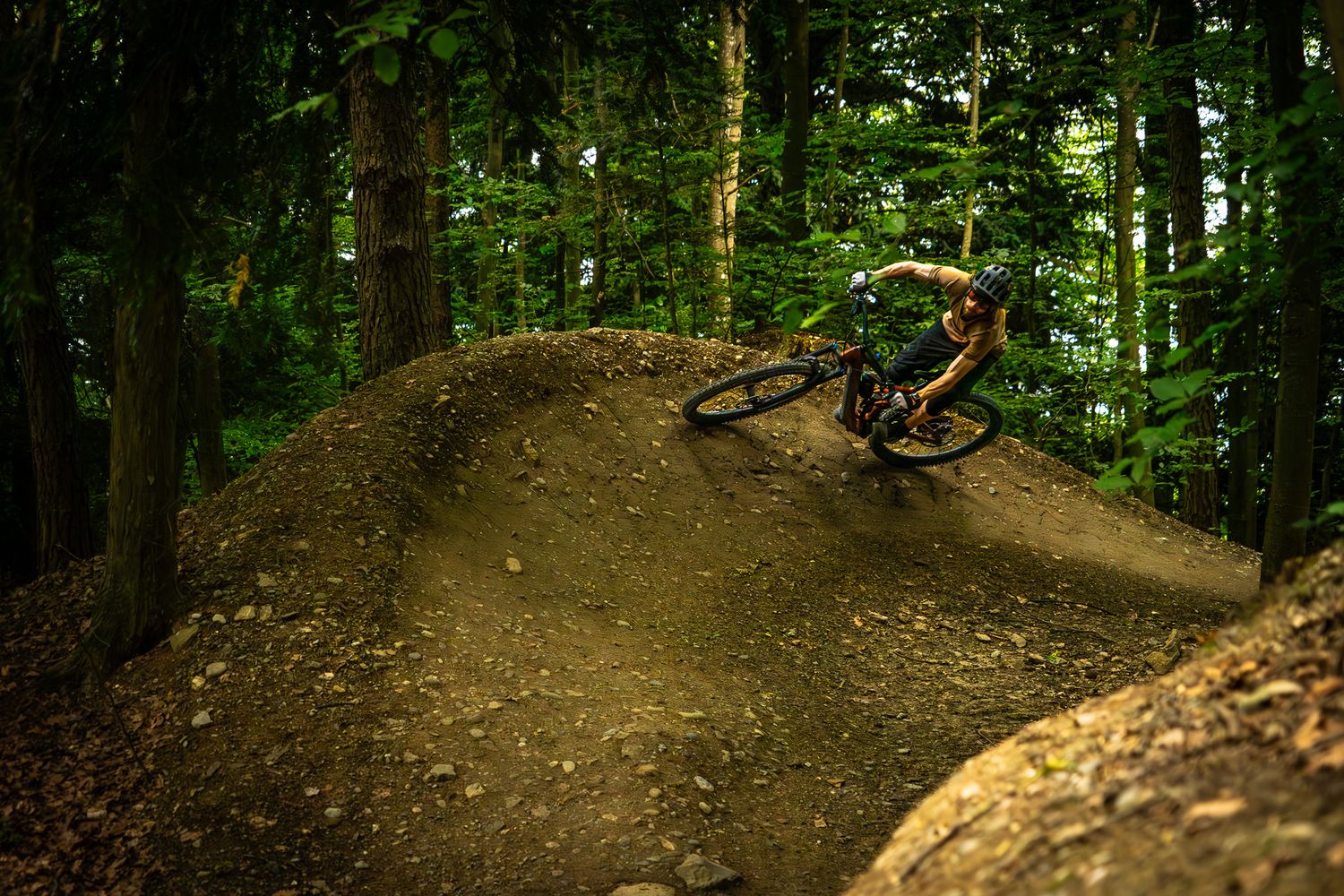 Mountain biker rides along curved dirt berm in dense woodland setting.