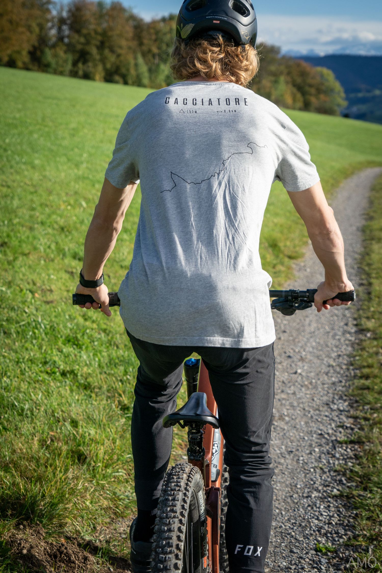 A cyclist rides along a gravel path through green alpine meadows with mountains in the distance.