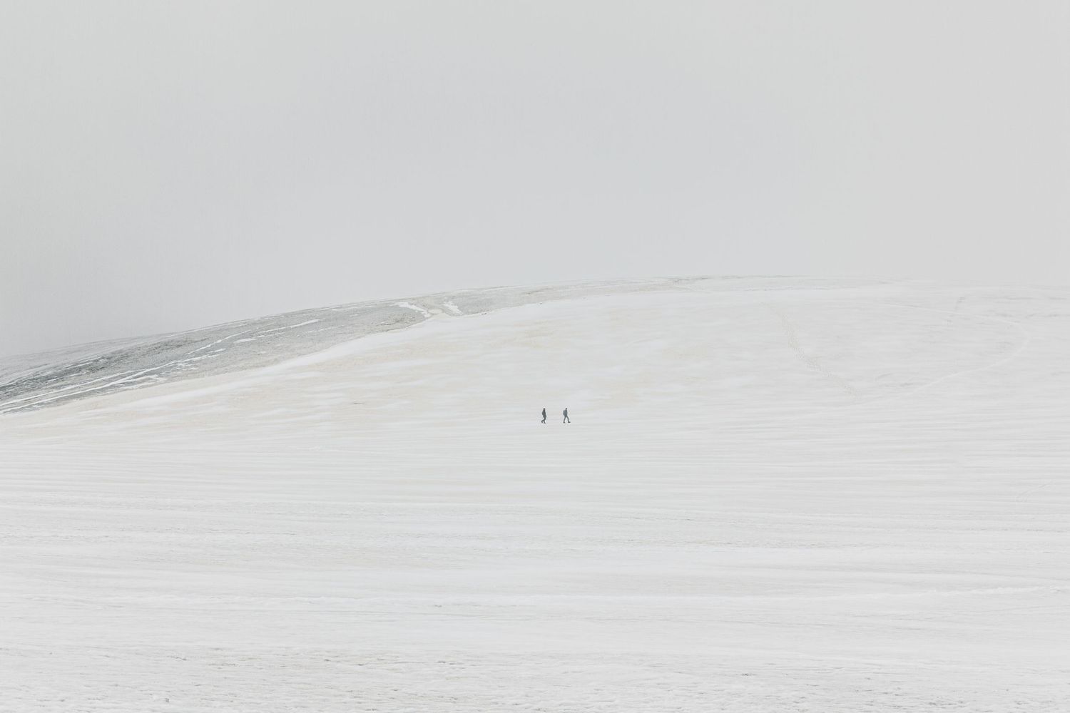 Minimalist winter landscape showing two tiny figures crossing a vast snowy slope.