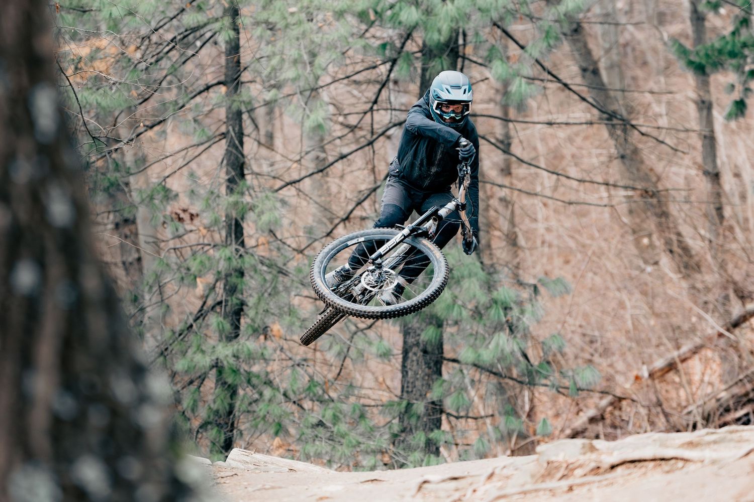 Mountain biker performing aerial jump with bike between trees in forest setting.