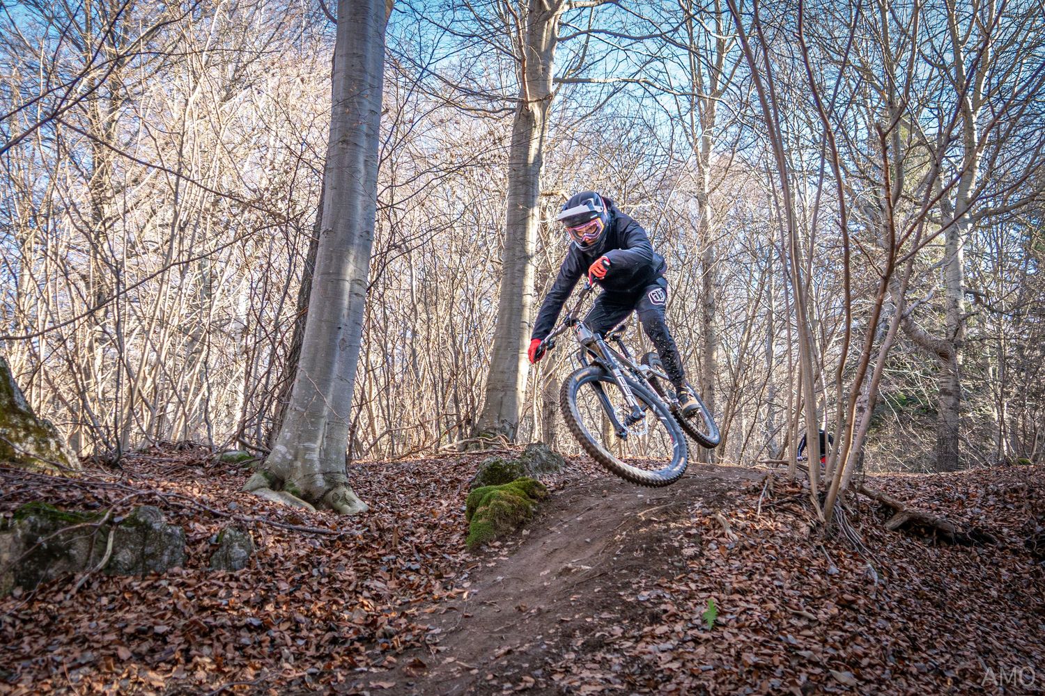 Mountain biker catching air off a dirt jump in a winter forest setting.