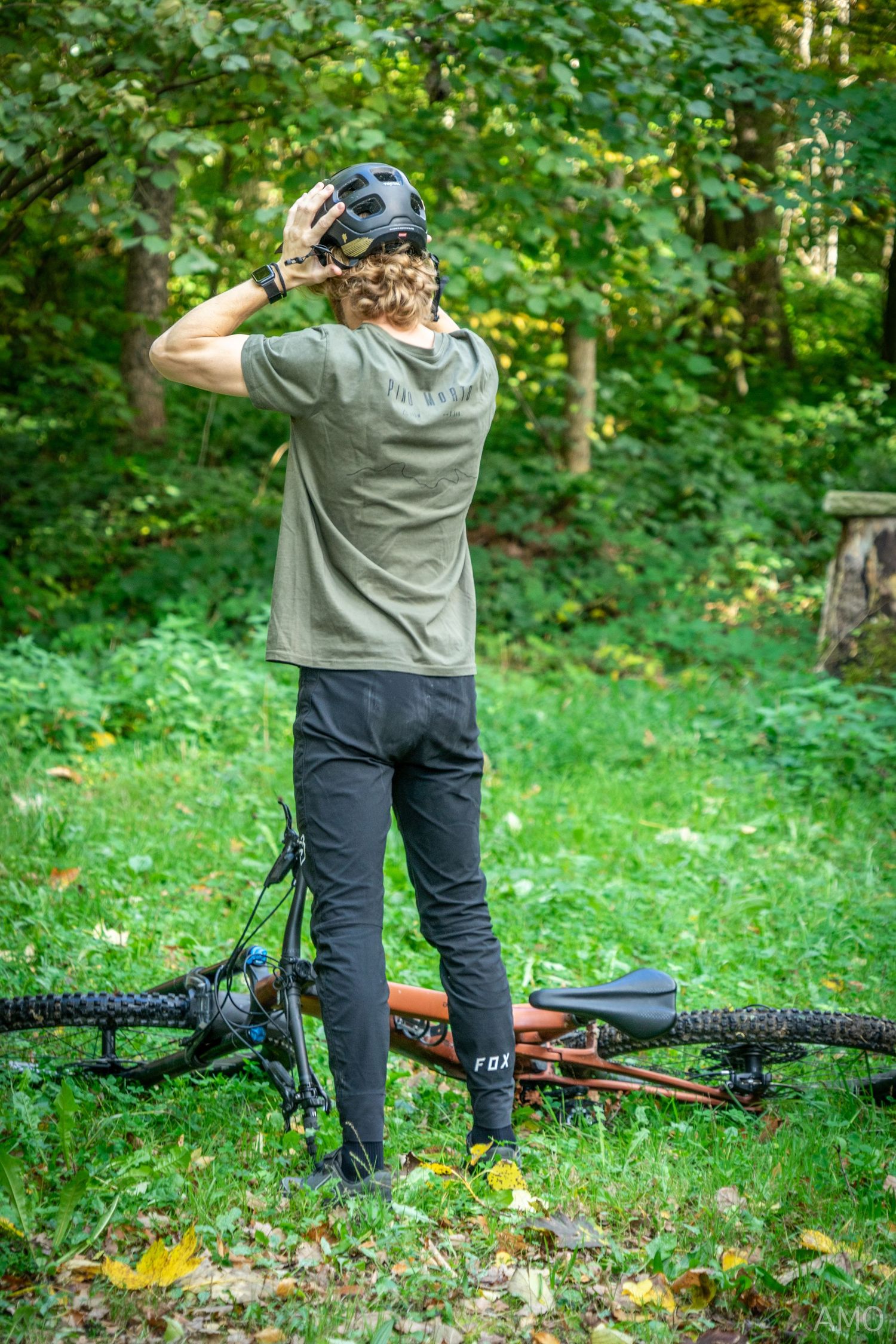 A cyclist takes a break near their mountain bike on a forest trail surrounded by green foliage.
