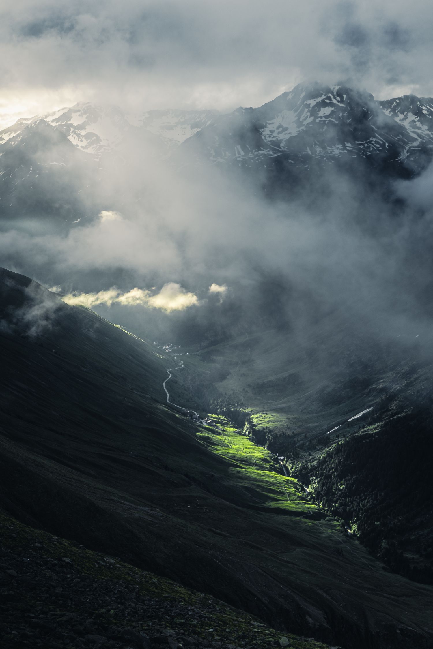 Misty mountain valley with dramatic sunlight illuminating a green patch of land below cloudy peaks.
