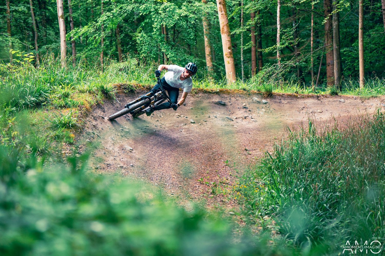 Mountain biker leans into a curved dirt trail turn surrounded by lush forest.