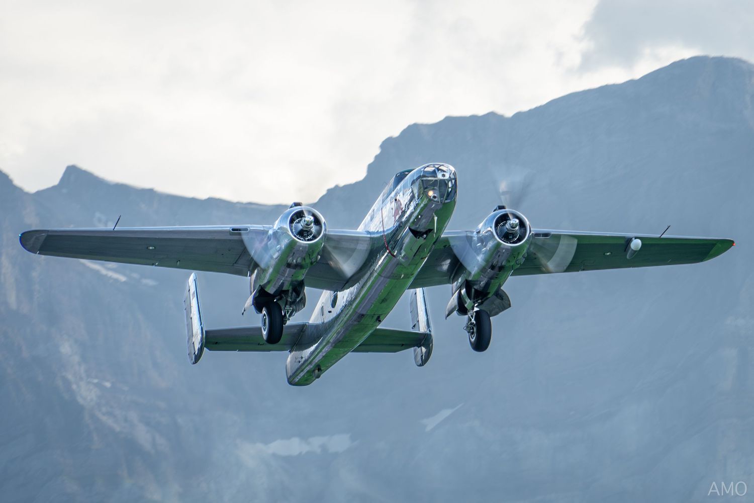 Vintage military aircraft with twin propellers flying against mountainous backdrop.
