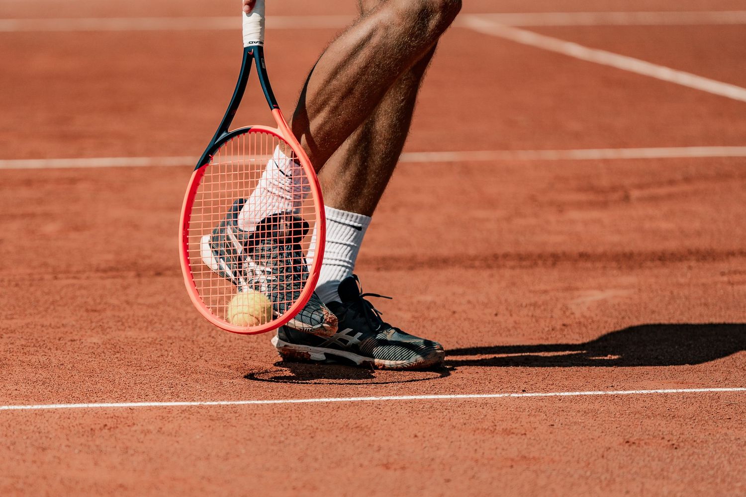 Tennis player practicing serves on a clay court with shadow cast on red surface.