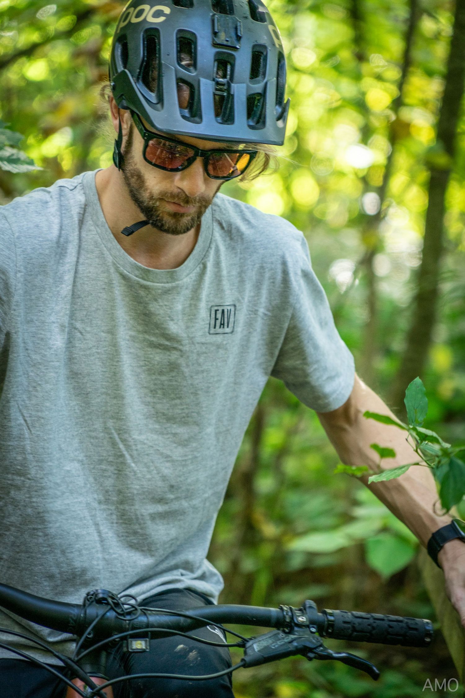 A cyclist in a grey t-shirt and helmet checks their gear while standing with their mountain bike in the woods.