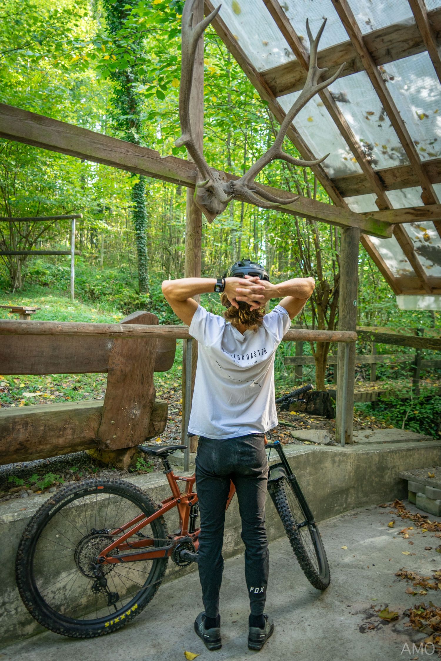 A mountain biker stretches under a wooden shelter in a lush forest setting with their orange bike nearby.