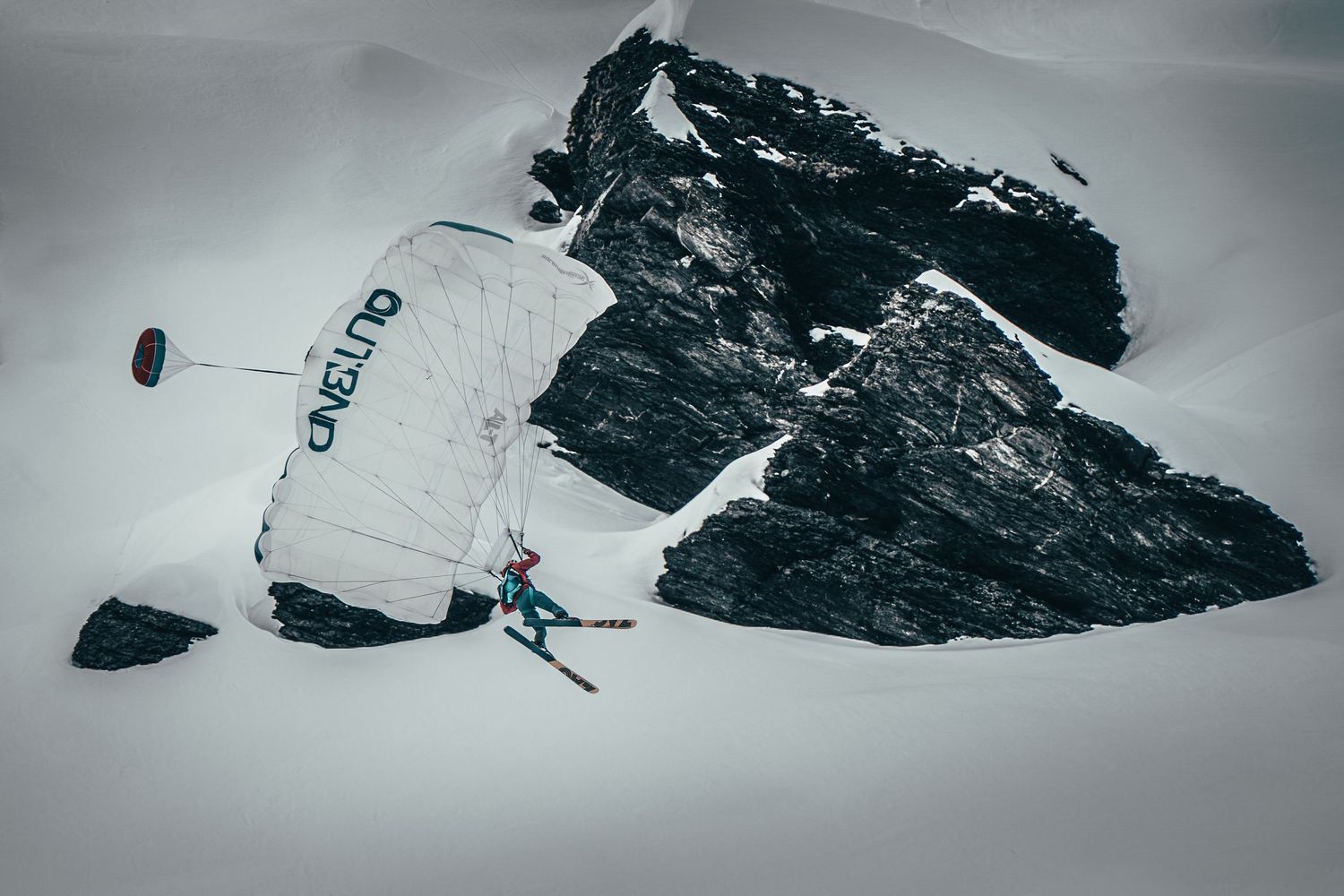 A skier performs a dramatic jump against a snowy mountain backdrop with a parachute visible.