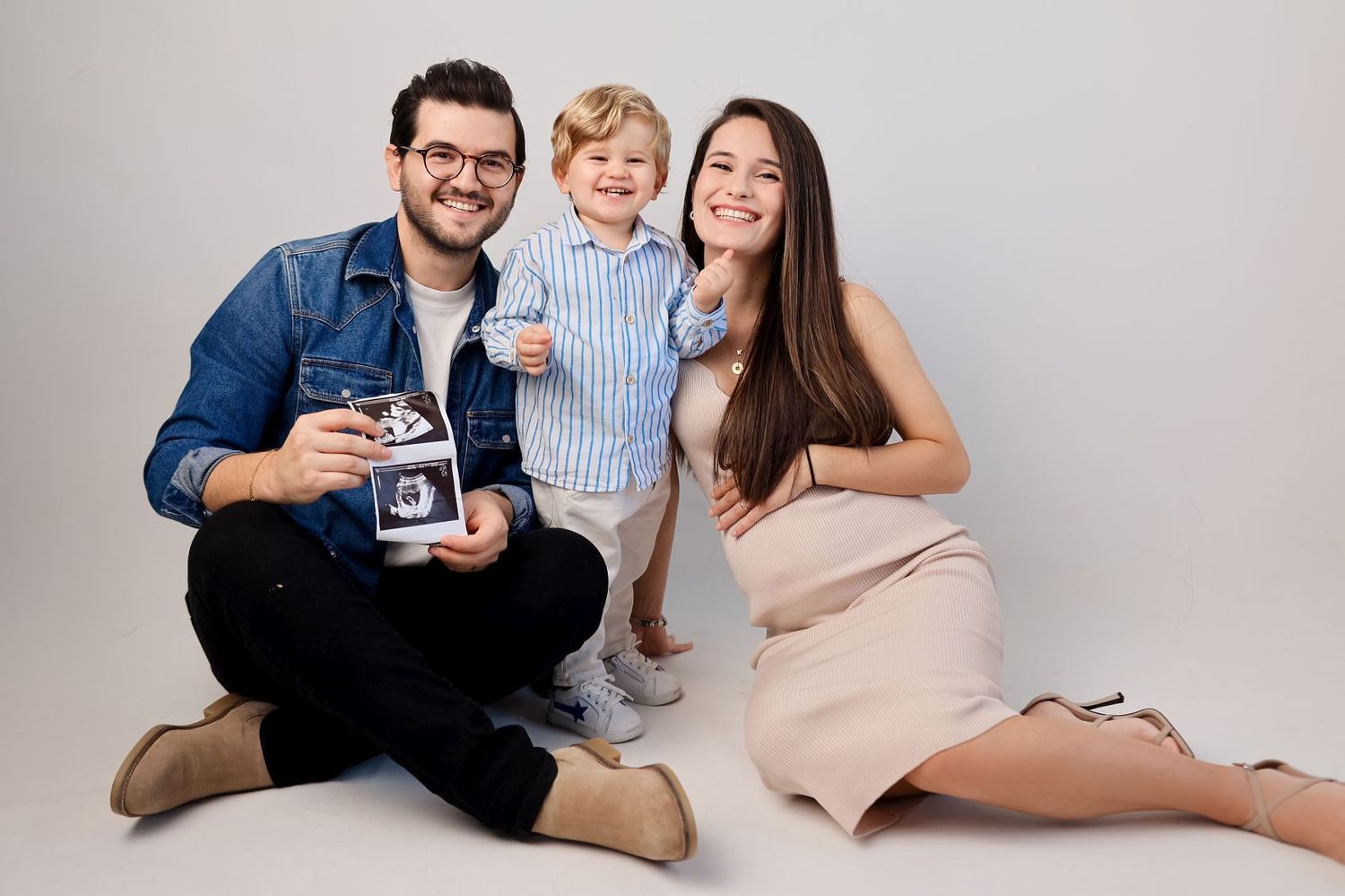 Family photoshoot with parents and child posing on white background with sonogram photos.
