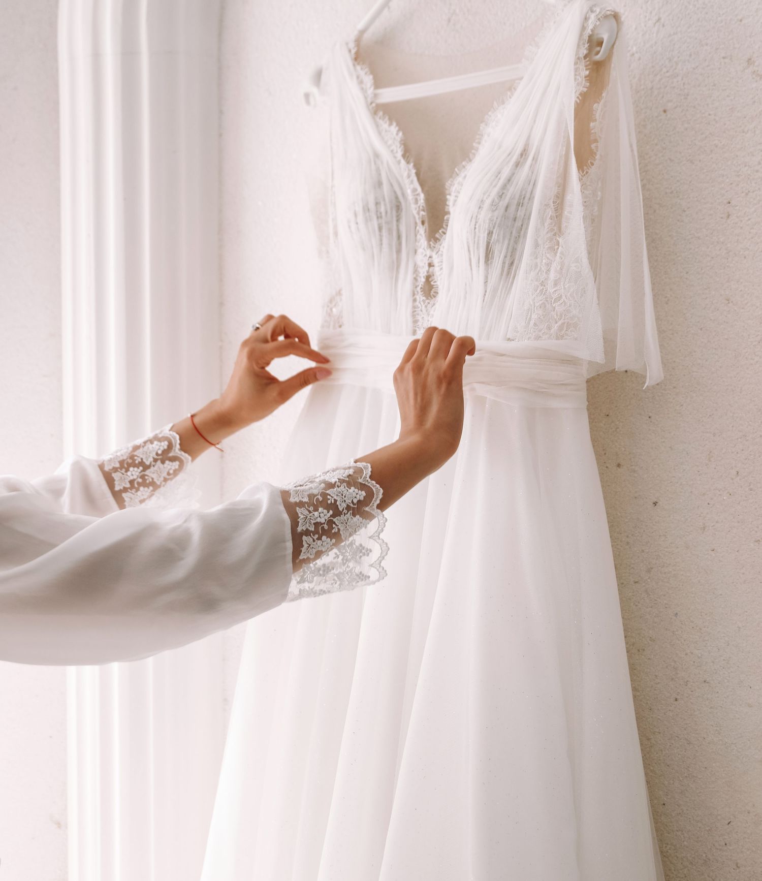 Delicate lace-sleeved hands adjusting an elegant white wedding dress on a hanger.