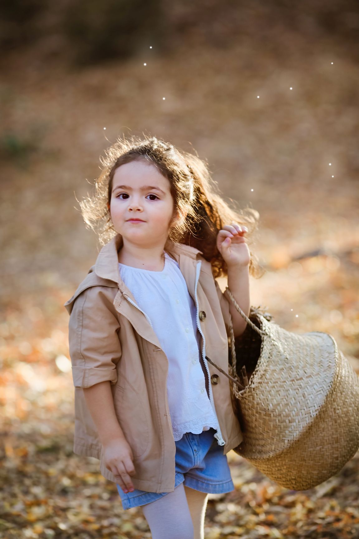 A child in a beige jacket holding a woven basket amid falling autumn leaves.