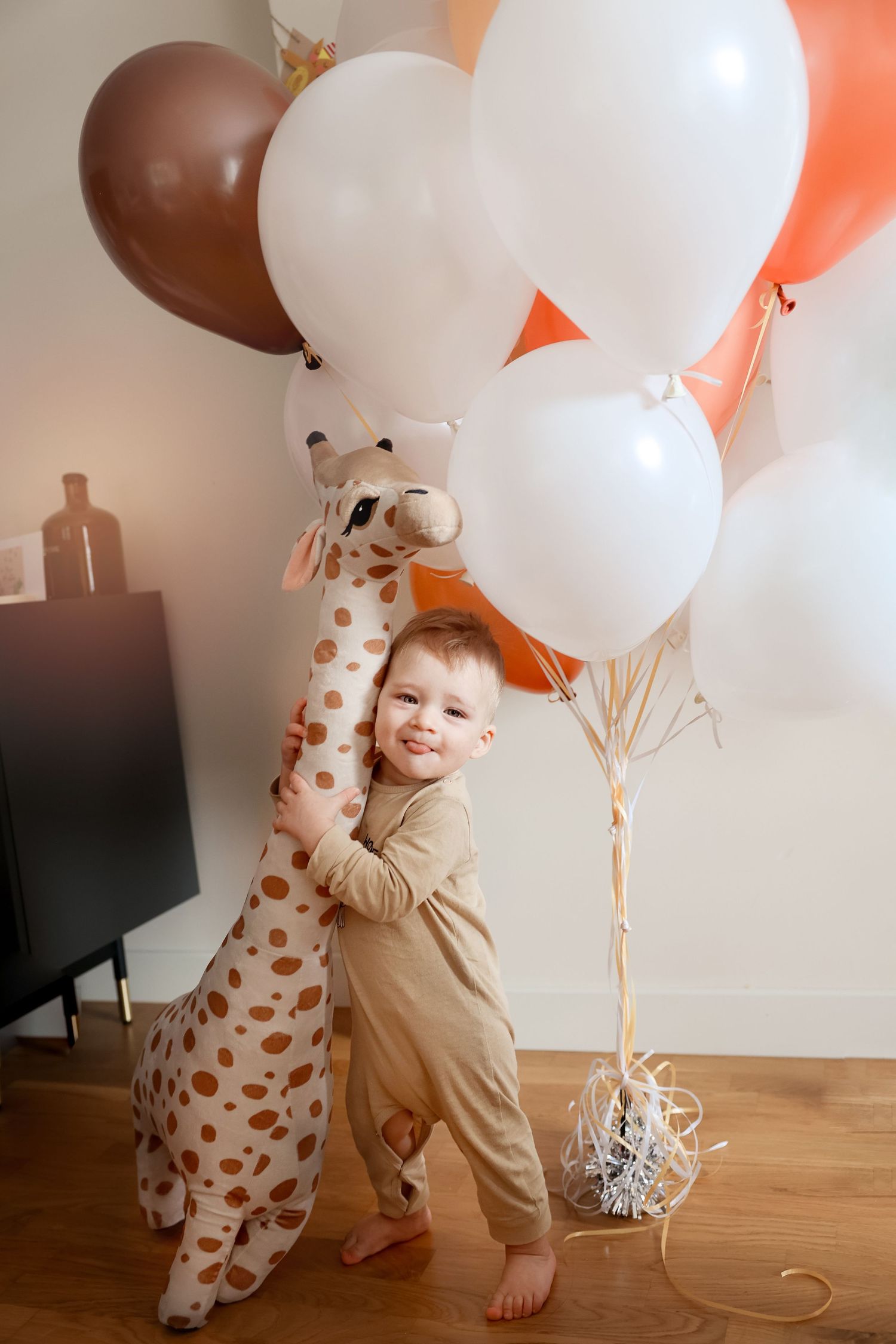 Small child hugging giraffe plush toy surrounded by peach and white balloons.
