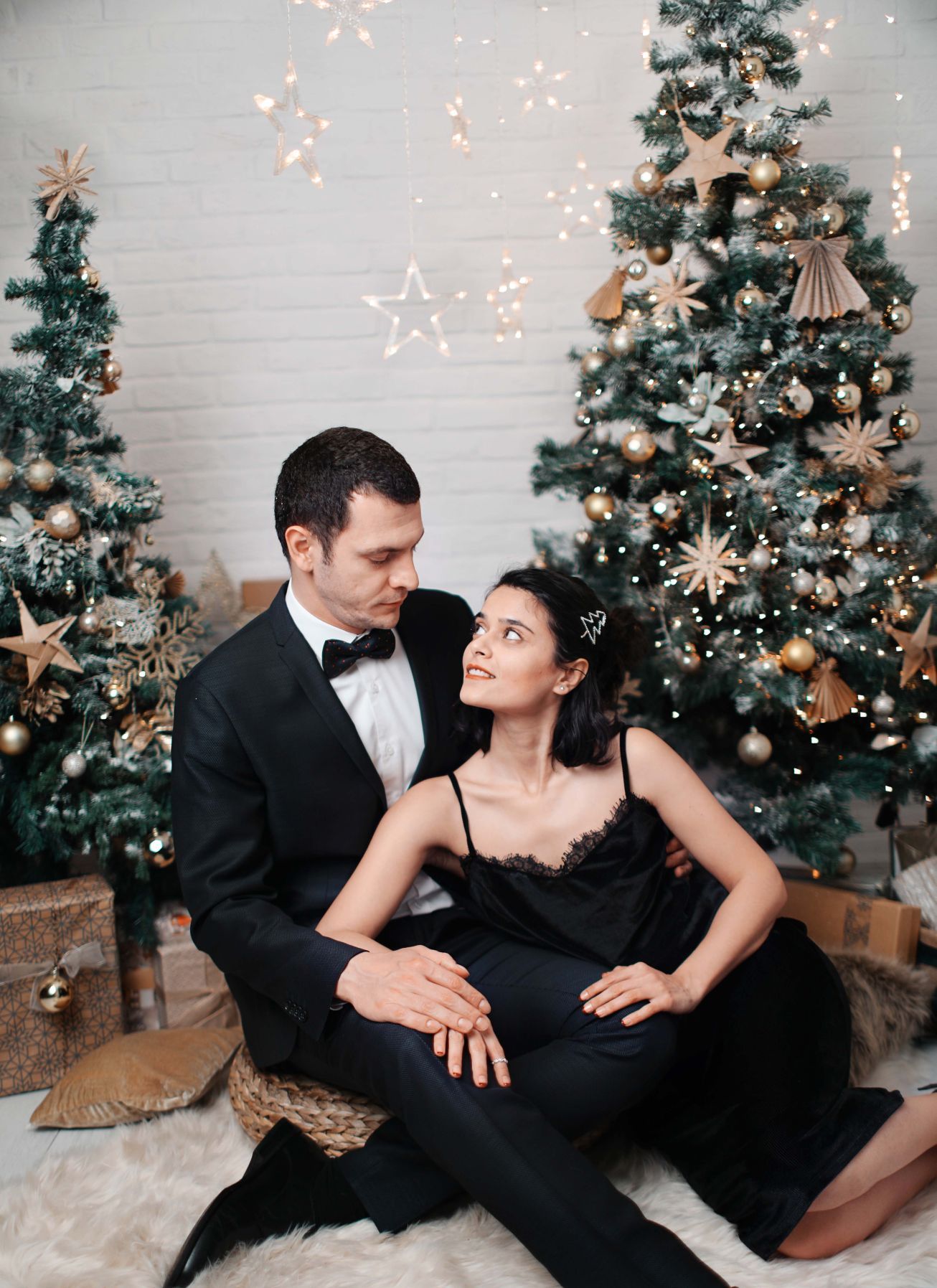 Elegant couple in formal attire poses in front of decorated Christmas trees with twinkling lights.