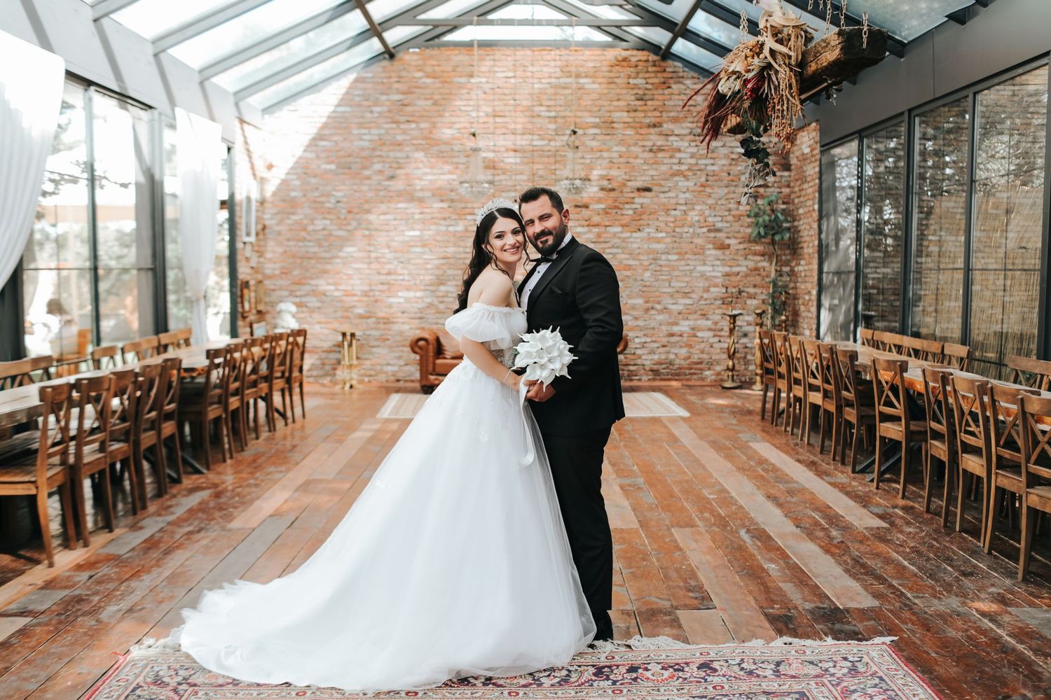 A wedding couple stands in an elegant venue with exposed brick walls and wooden chairs.