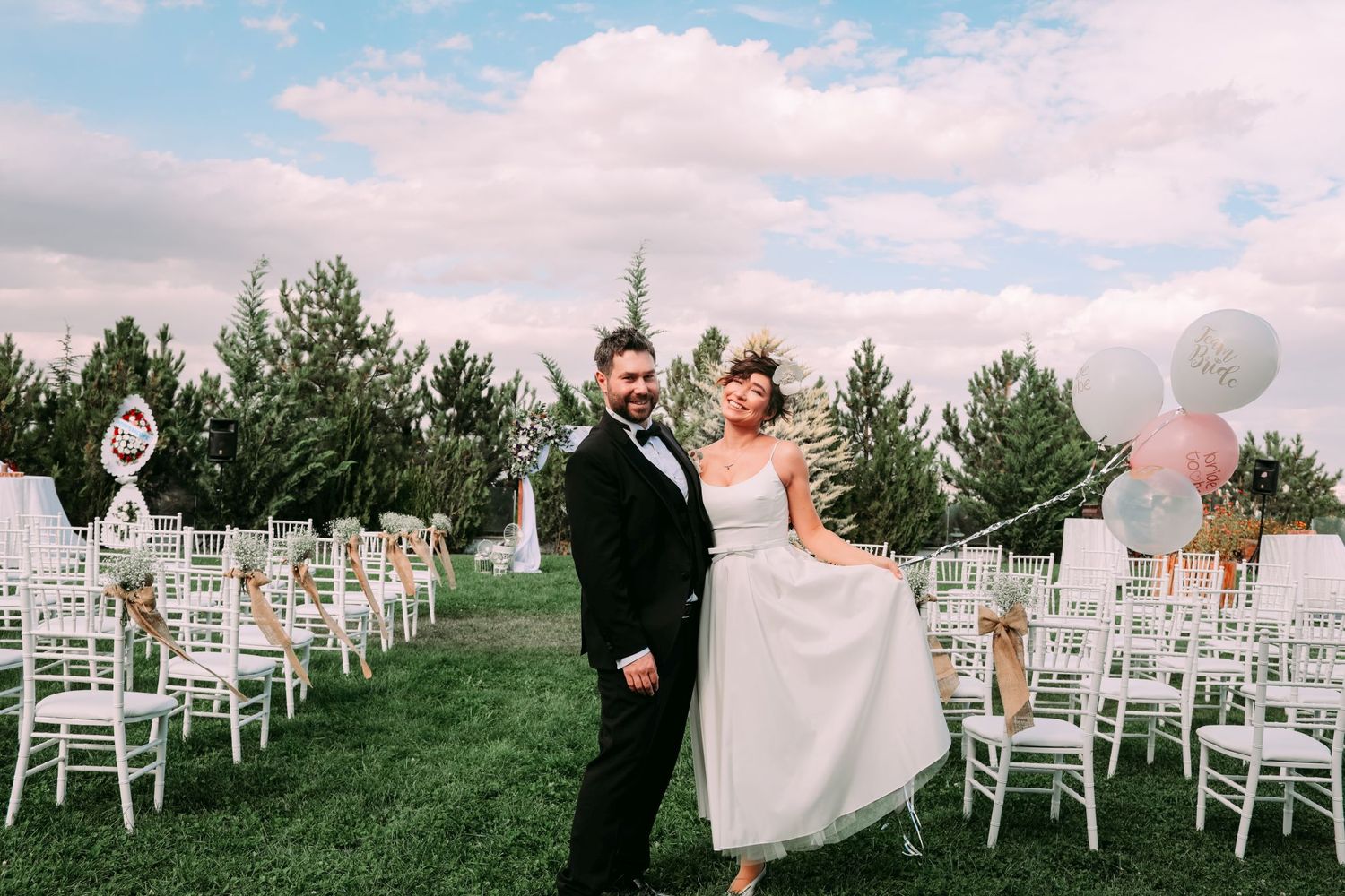 A festive outdoor wedding ceremony setup with white chairs and pink balloons under blue sky.