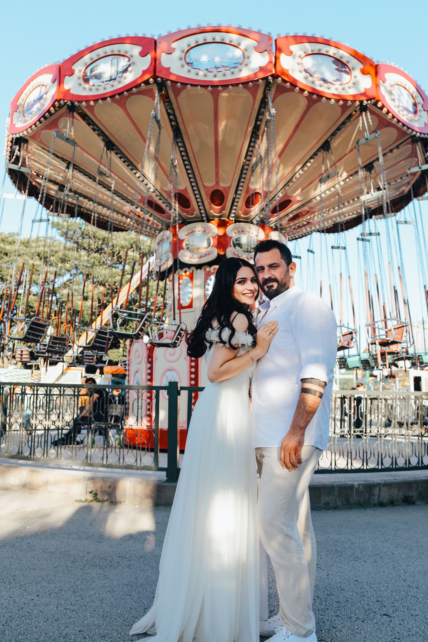 A couple in white attire poses in front of a vintage carousel at an amusement park.